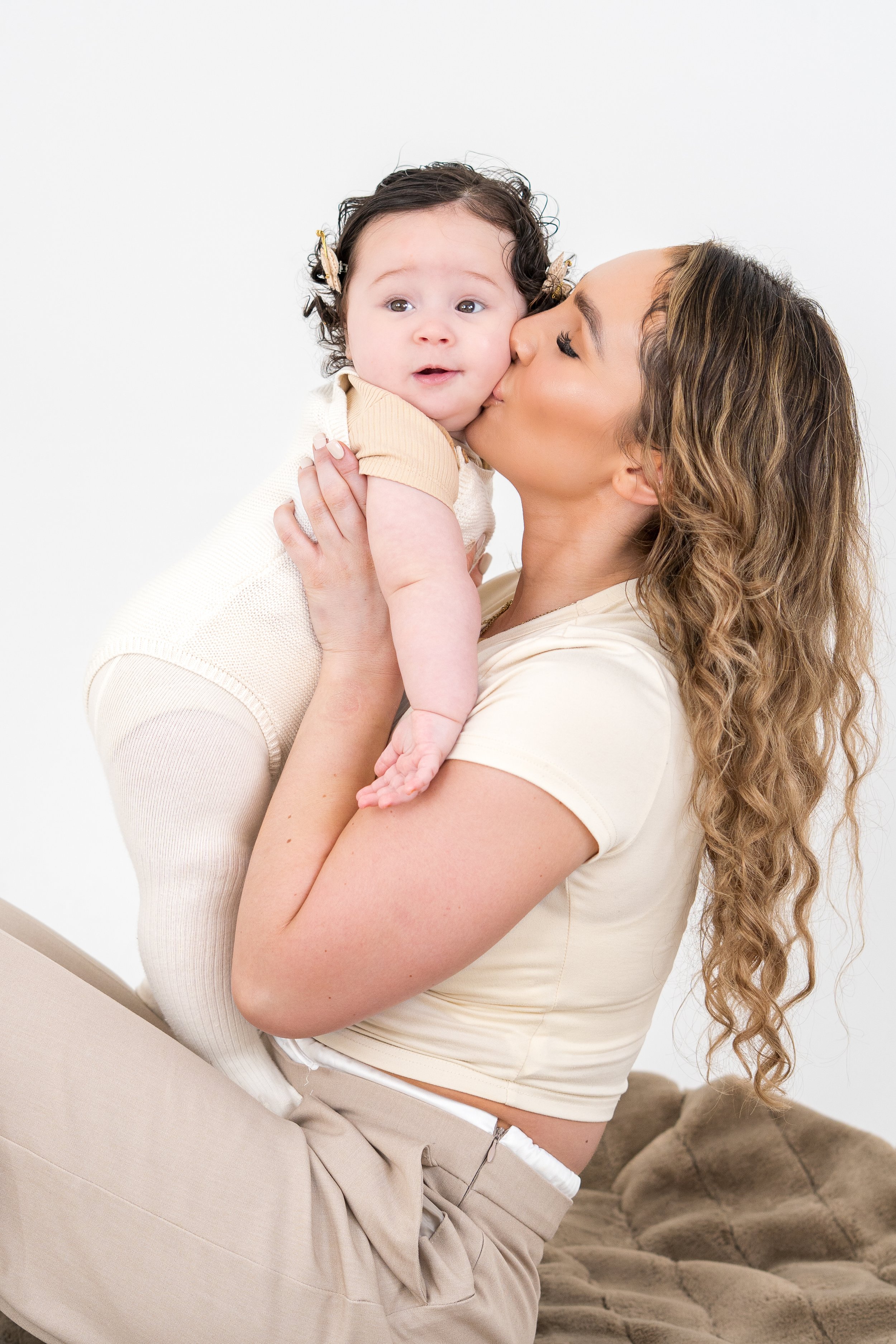 A woman with curly hair kisses a young girl on the cheek, who has dark curly hair and is wearing a light beige outfit, against a plain white background.