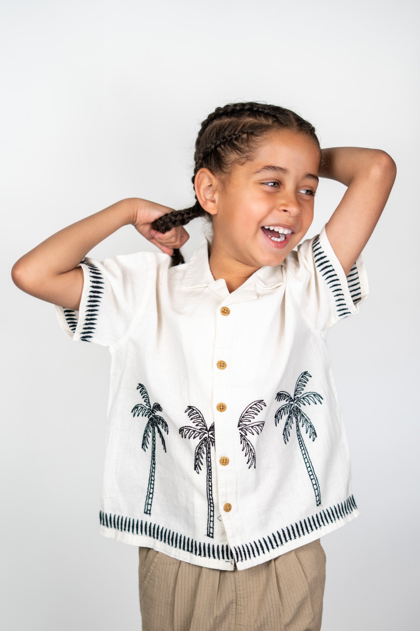 A young girl with braided hair smiling and adjusting her hair, wearing a white shirt with palm tree and striped details, standing against a plain white background.