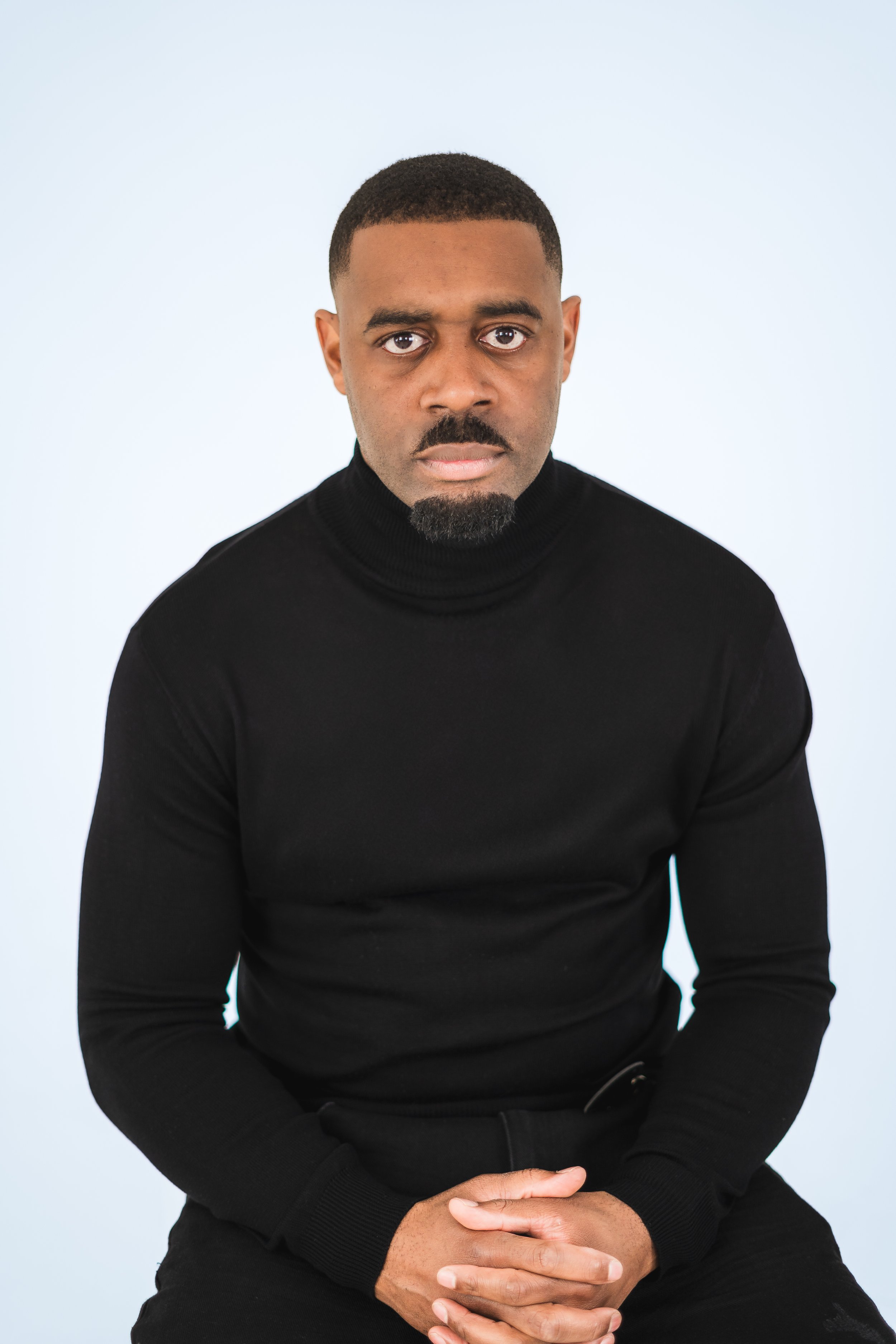 A man with a short haircut and a beard, wearing a black turtleneck, looking serious, seated against a plain light background.