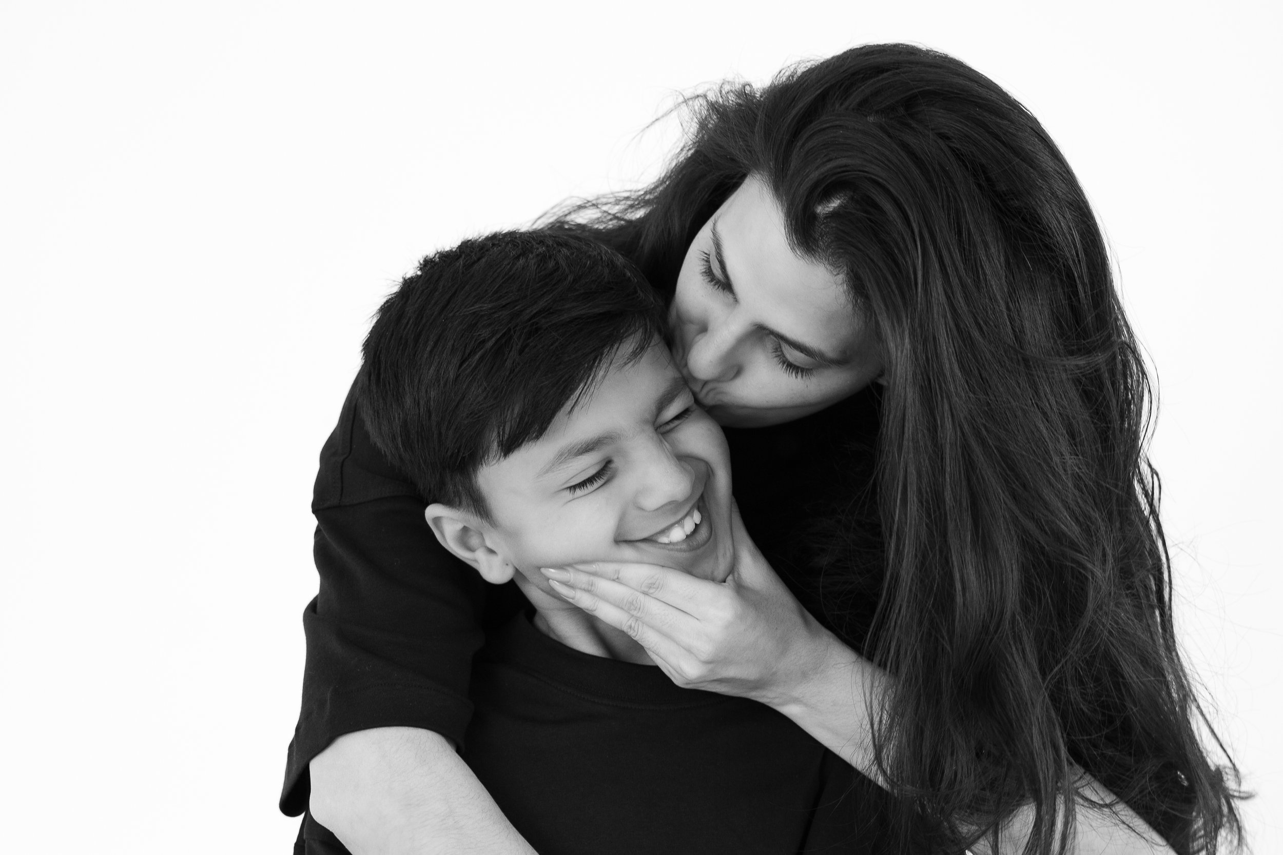 A woman hugging a smiling young boy, kissing him on the forehead in a black and white photo.