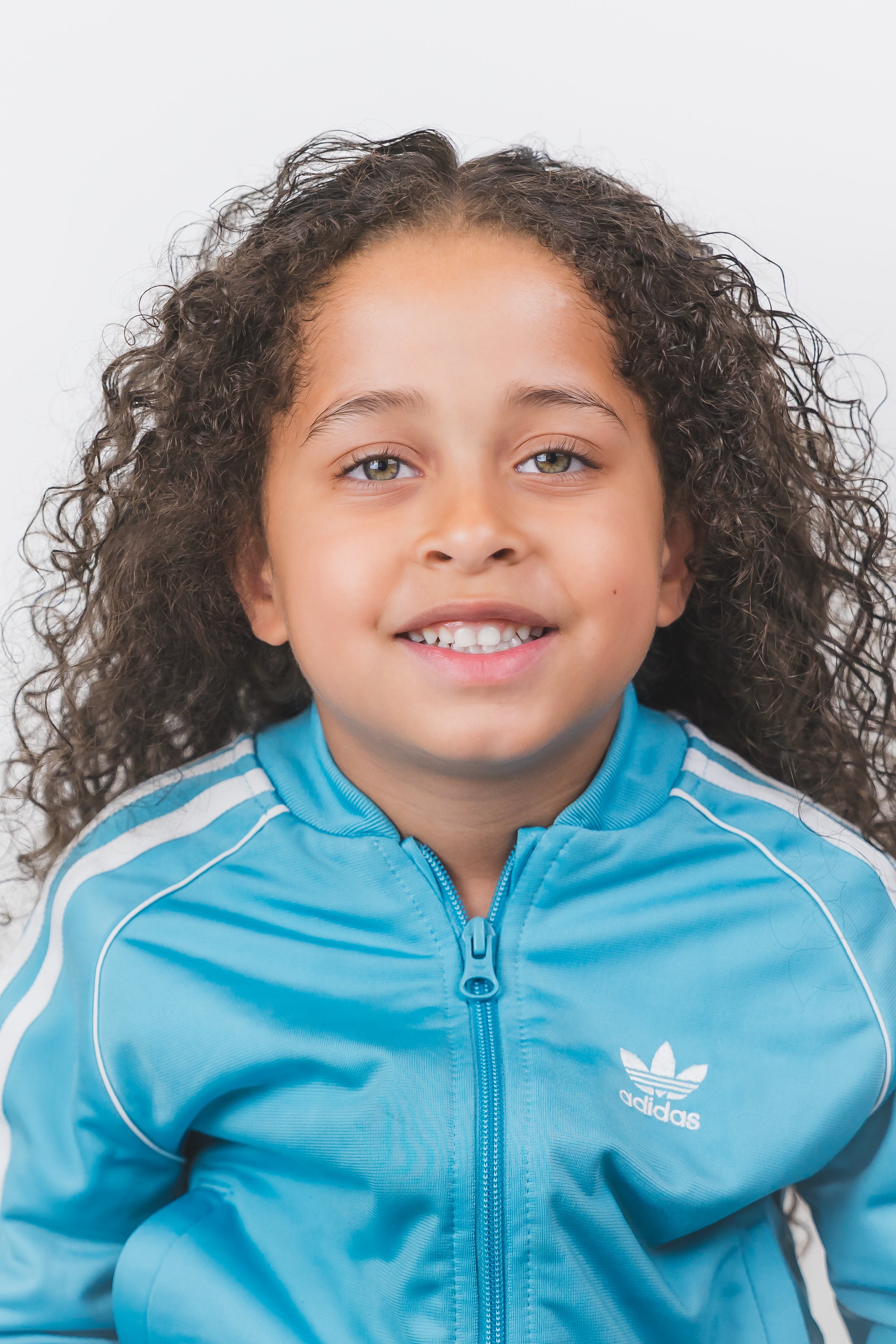 Young girl with curly hair smiling, wearing a blue Adidas jacket, against a white background.