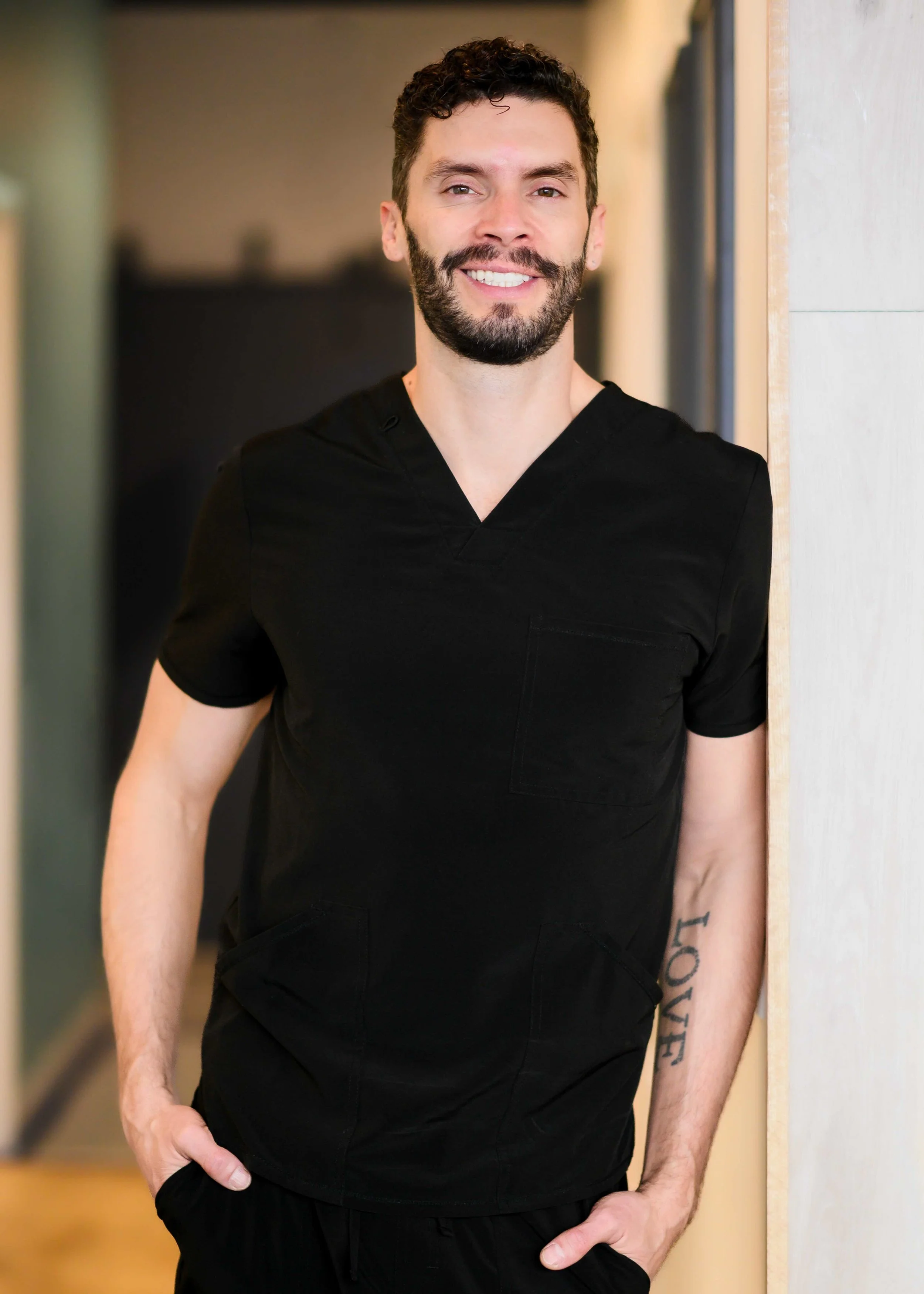 A smiling man with curly dark hair, beard, and mustache wearing black medical scrubs, standing indoors near a wall.
