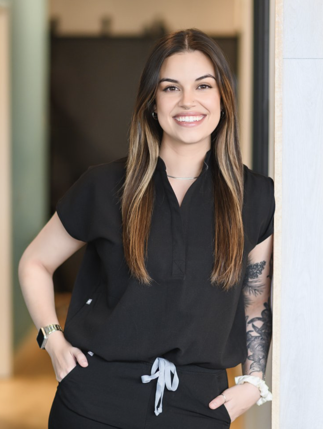 A young woman with long brown hair and a tattoo on her left arm, smiling and standing indoors near a doorway, wearing a black polo shirt and black pants with a gray tie belt.