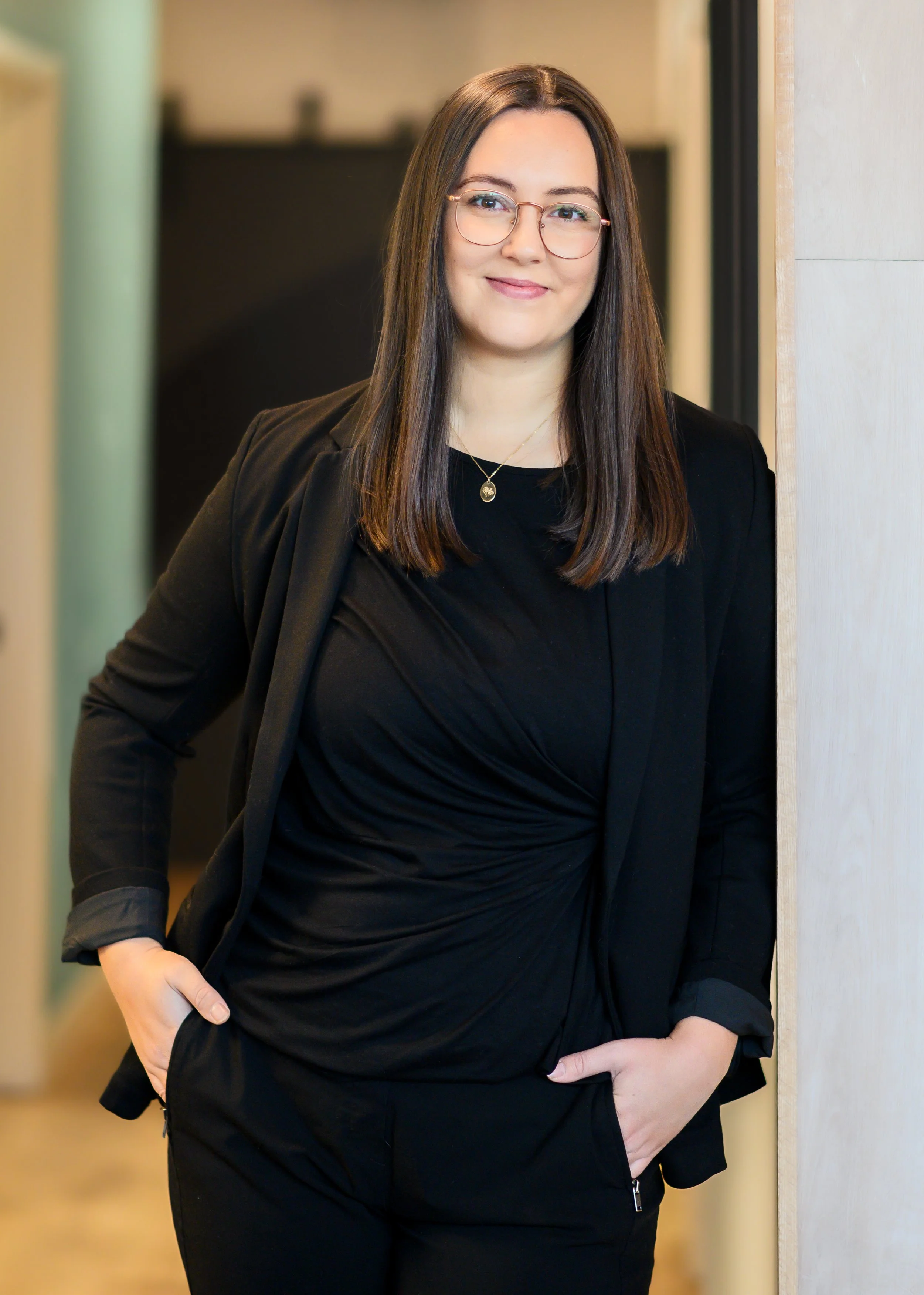 A woman with shoulder-length brown hair, wearing glasses and a black blazer, standing indoors with her hands in her pockets, smiling at the camera.