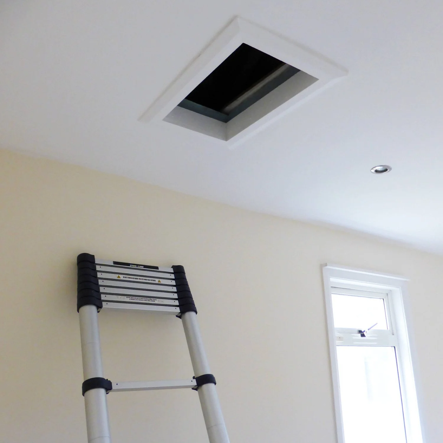 View of a room's ceiling with a ceiling vent, small window, and a folding ladder leaning against the wall.