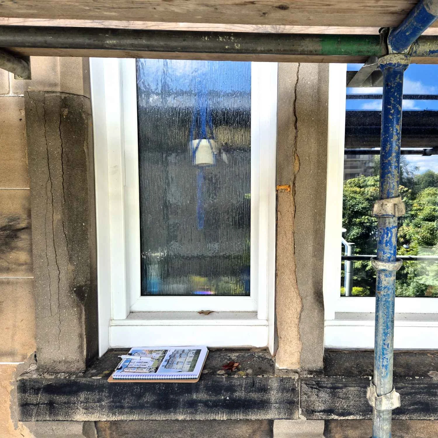Photo of a window on a building with scaffolding on the right side. A clip board is on the window ledge. The surrounding wall has visible cracks and is in the process of construction or renovation.