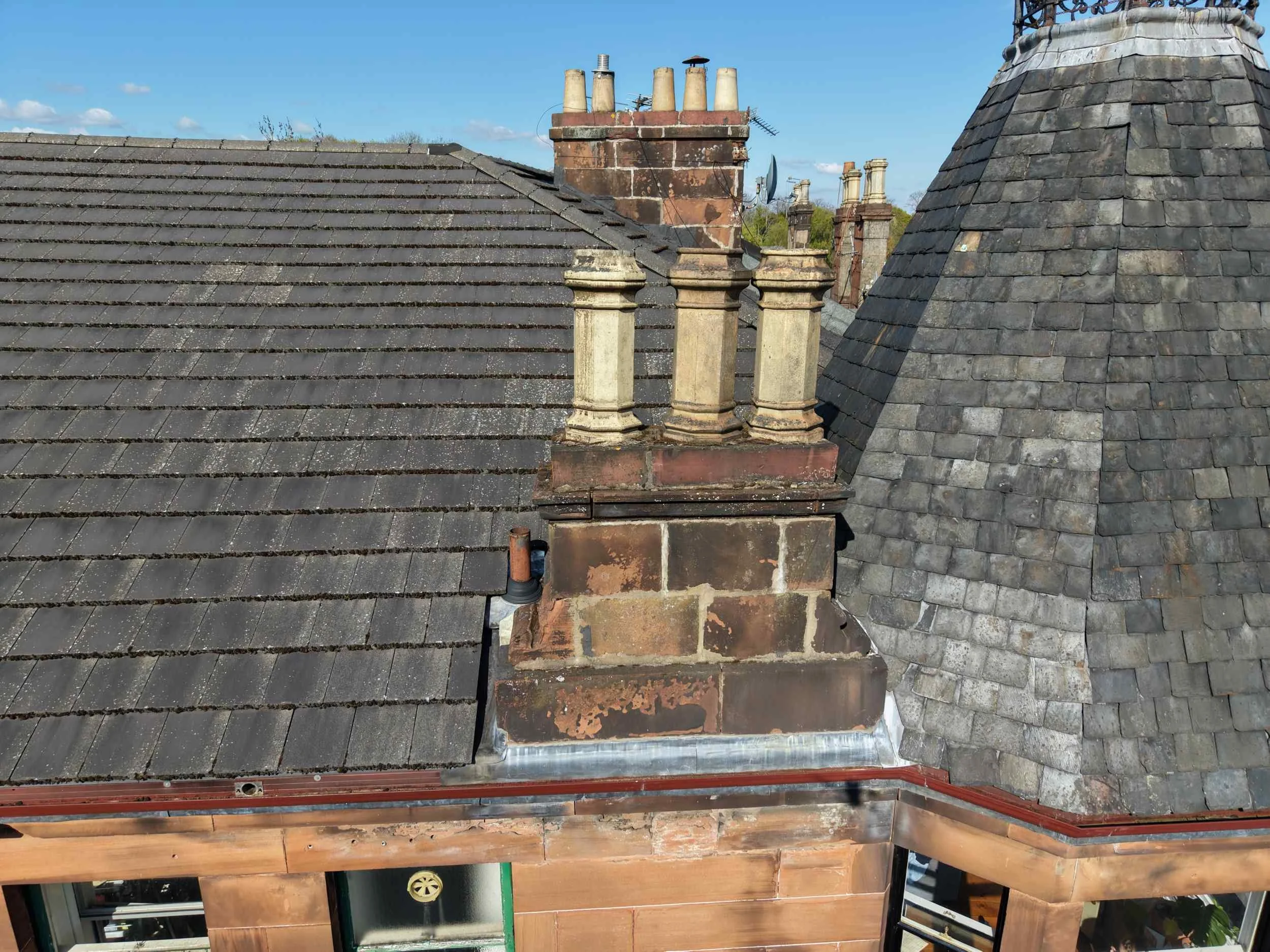 Photo of a tenement roof with multiple chimneys and grey cement tiles, some with moss and weathering, under a clear blue sky.