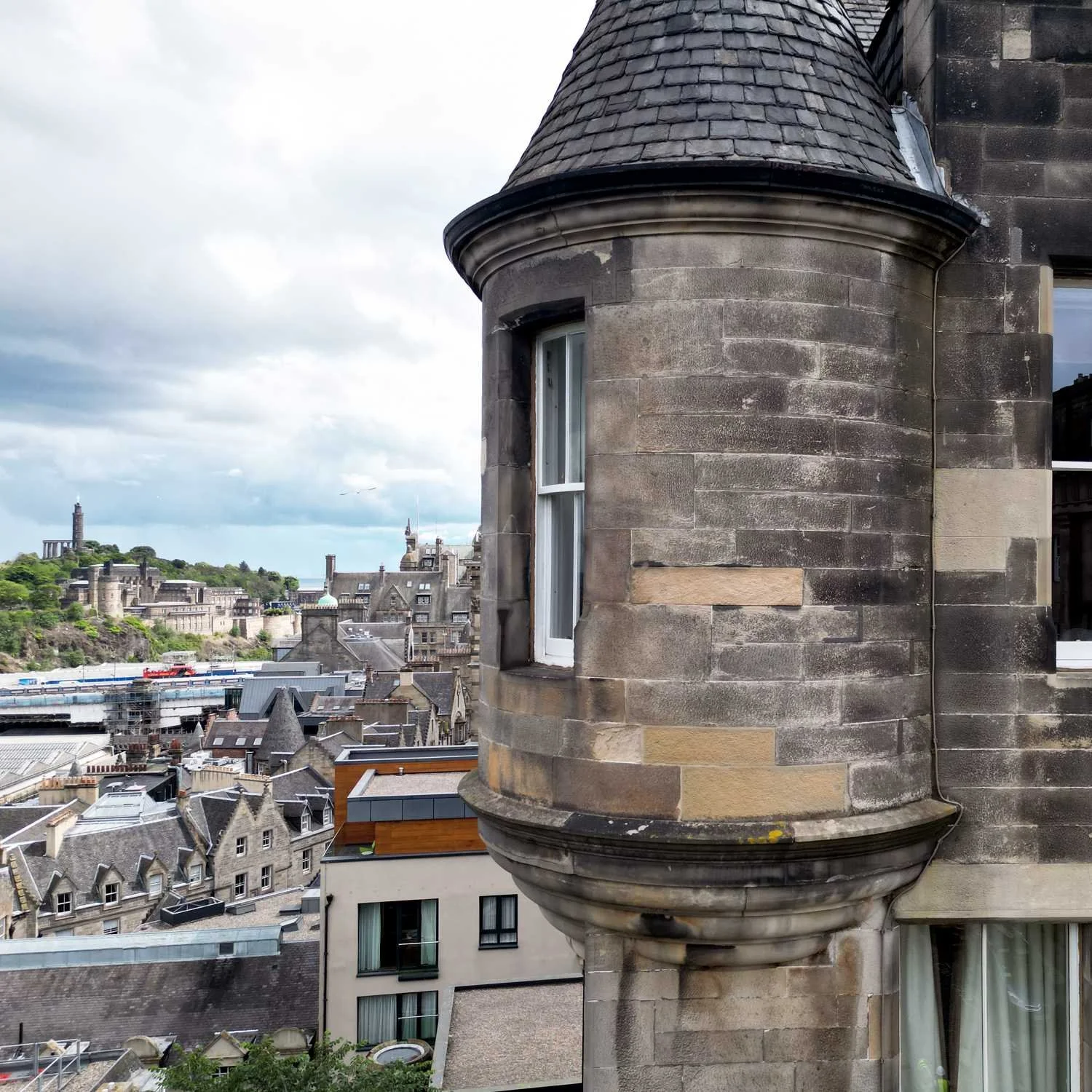 A view of a city skyline with historic buildings, green trees, and a hill in the distance, seen from a high-rise building with stone walls, windows, and a turret-like architectural feature.