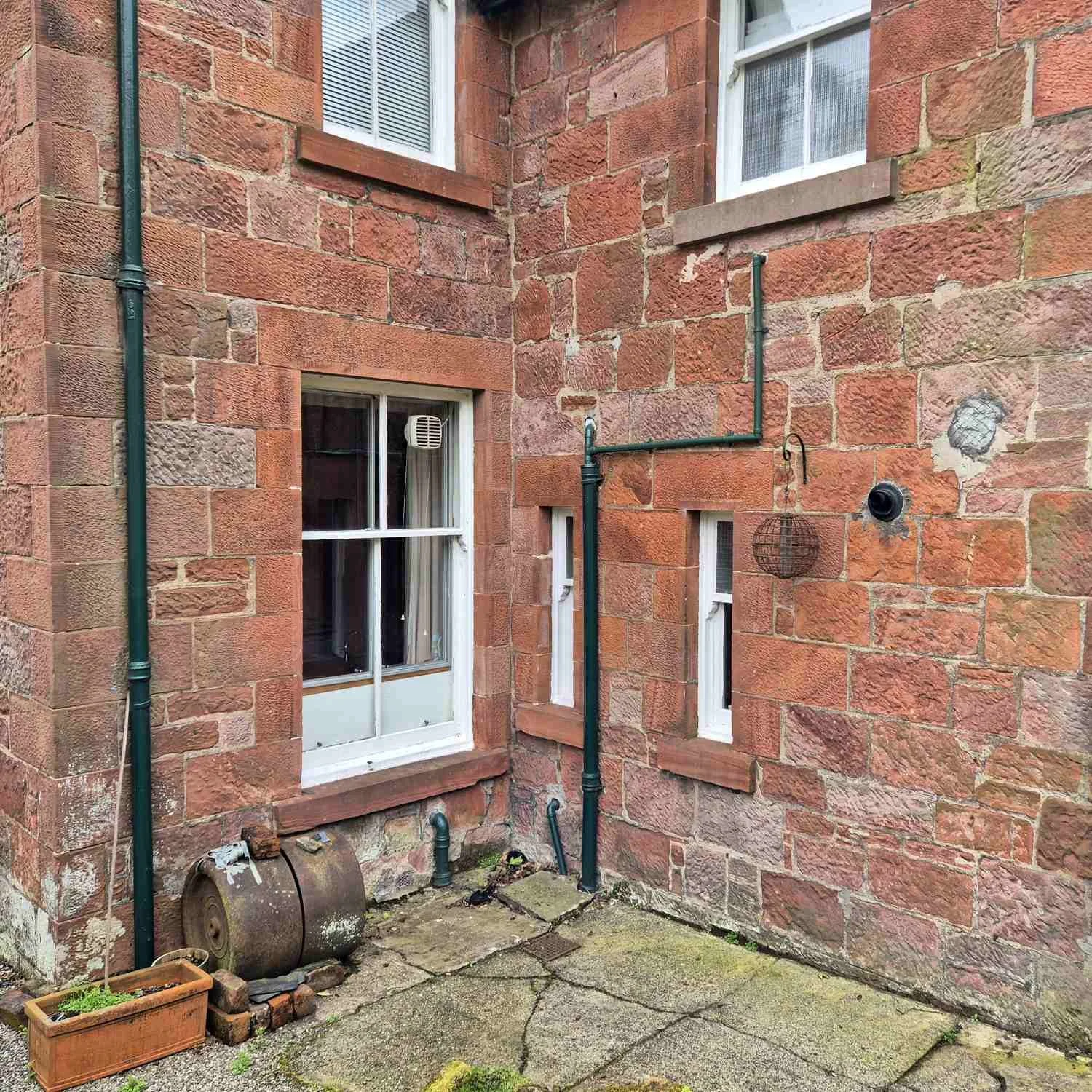 Backyard corner of a stone house with two windows, a door, green gutters, and various pipes. The yard has a concrete patio with moss and cracks, an empty gardening barrel, and a small flower bed with a plastic container.