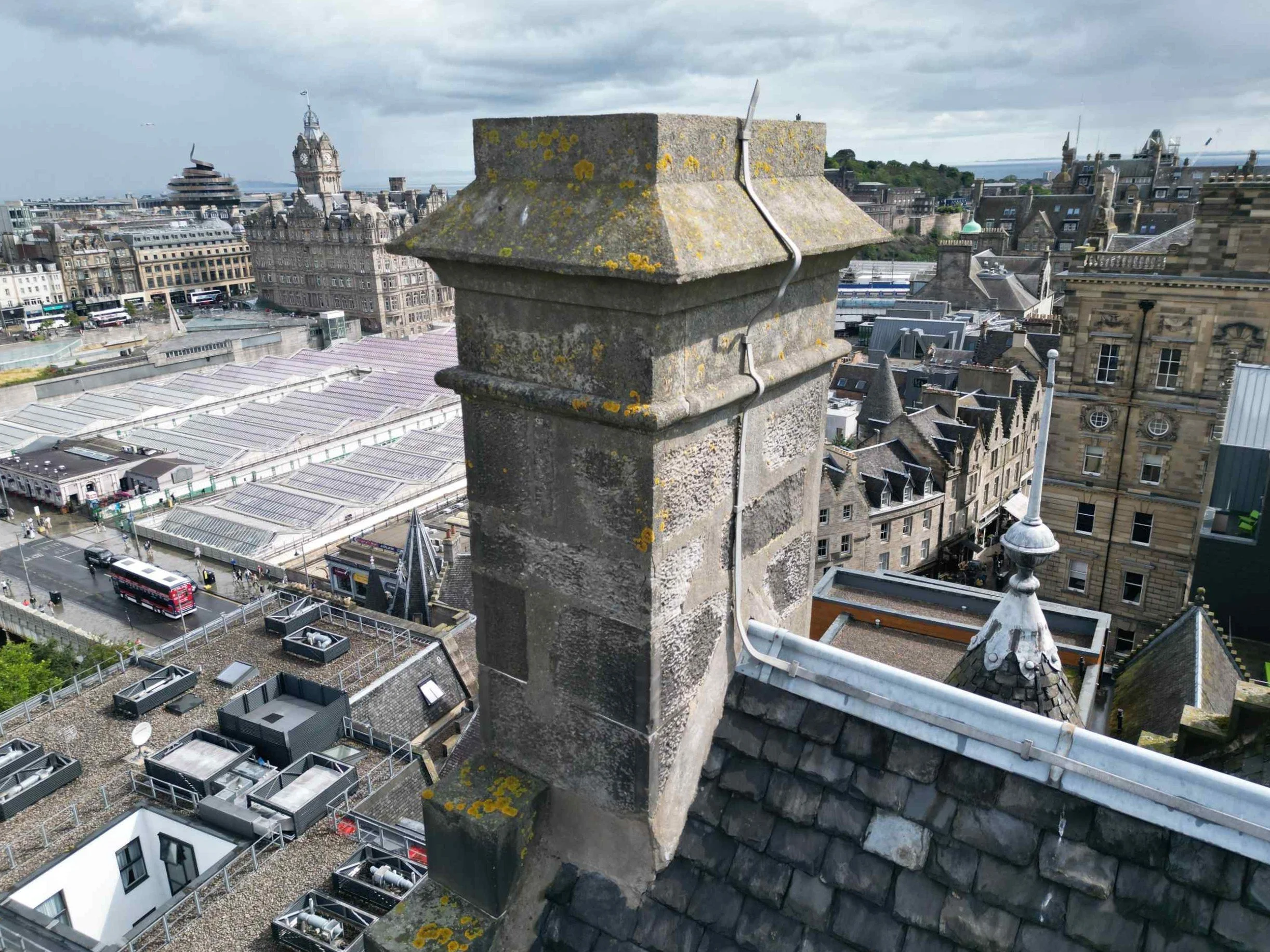 Close-up of a stone chimney stack on top of a historic building in a cityscape with an overcast sky, other old and modern buildings, and railway station below.