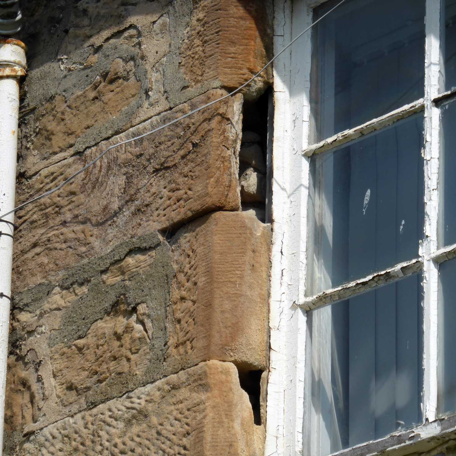 Close-up of a stone building wall with a window. The wall is made of large, uneven stones, some with textured surfaces. The window has a wooden frame painted white, with multiple small panes separated by white muntins, and the paint is peeling.