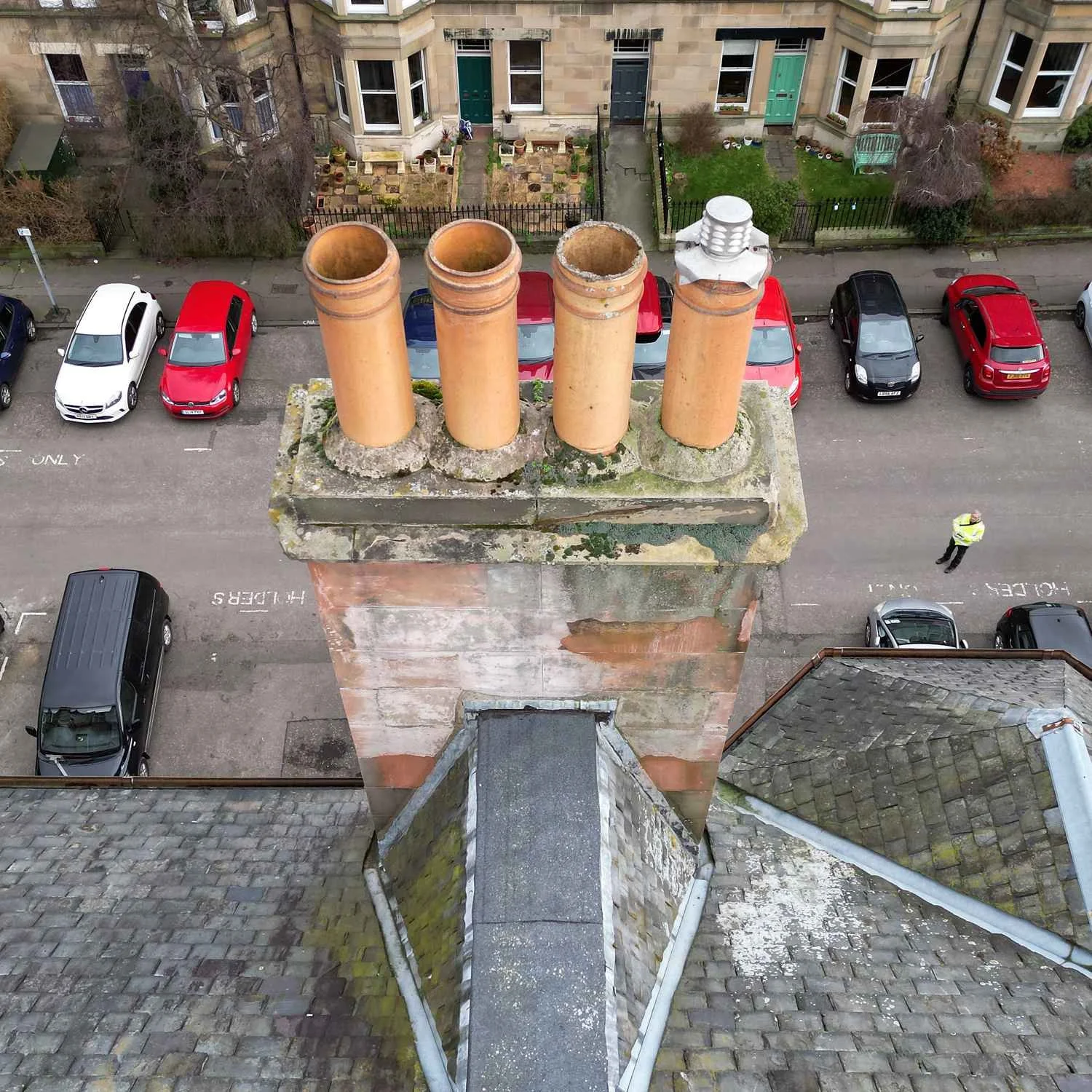 View from above of a chimney with four pots on top. Cars are parked on the street below, with houses and gardens in the background.