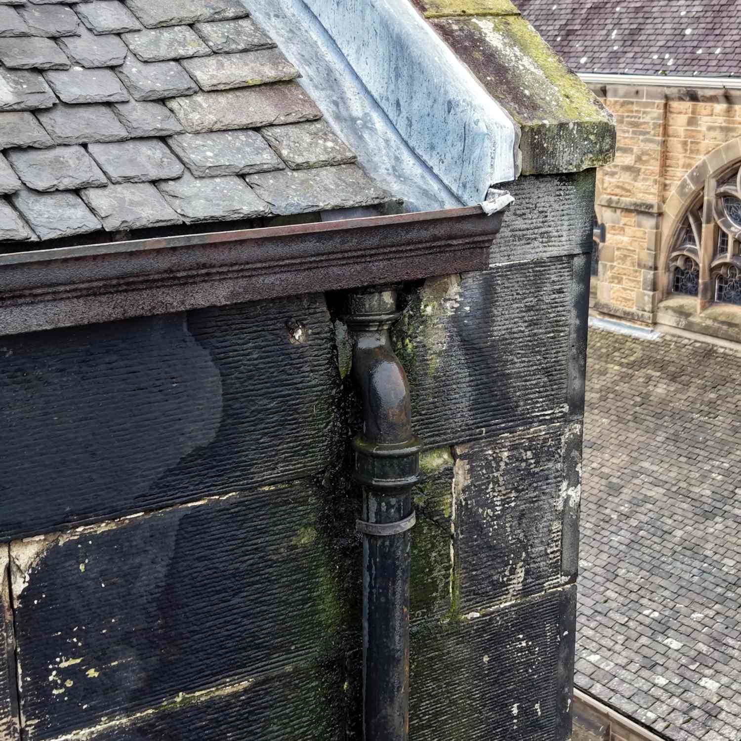 Close-up of a corner of a old stone building with a black drainpipe and a sloped grey slate roof, and a church or cathedral with stained glass windows in the background.