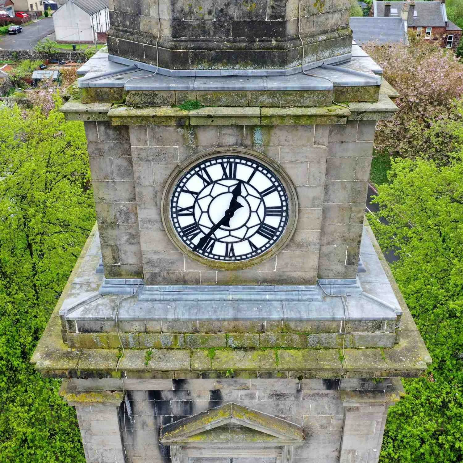 A clock tower with a black and white clock face showing the time as 12:37, surrounded by green trees and residential buildings.