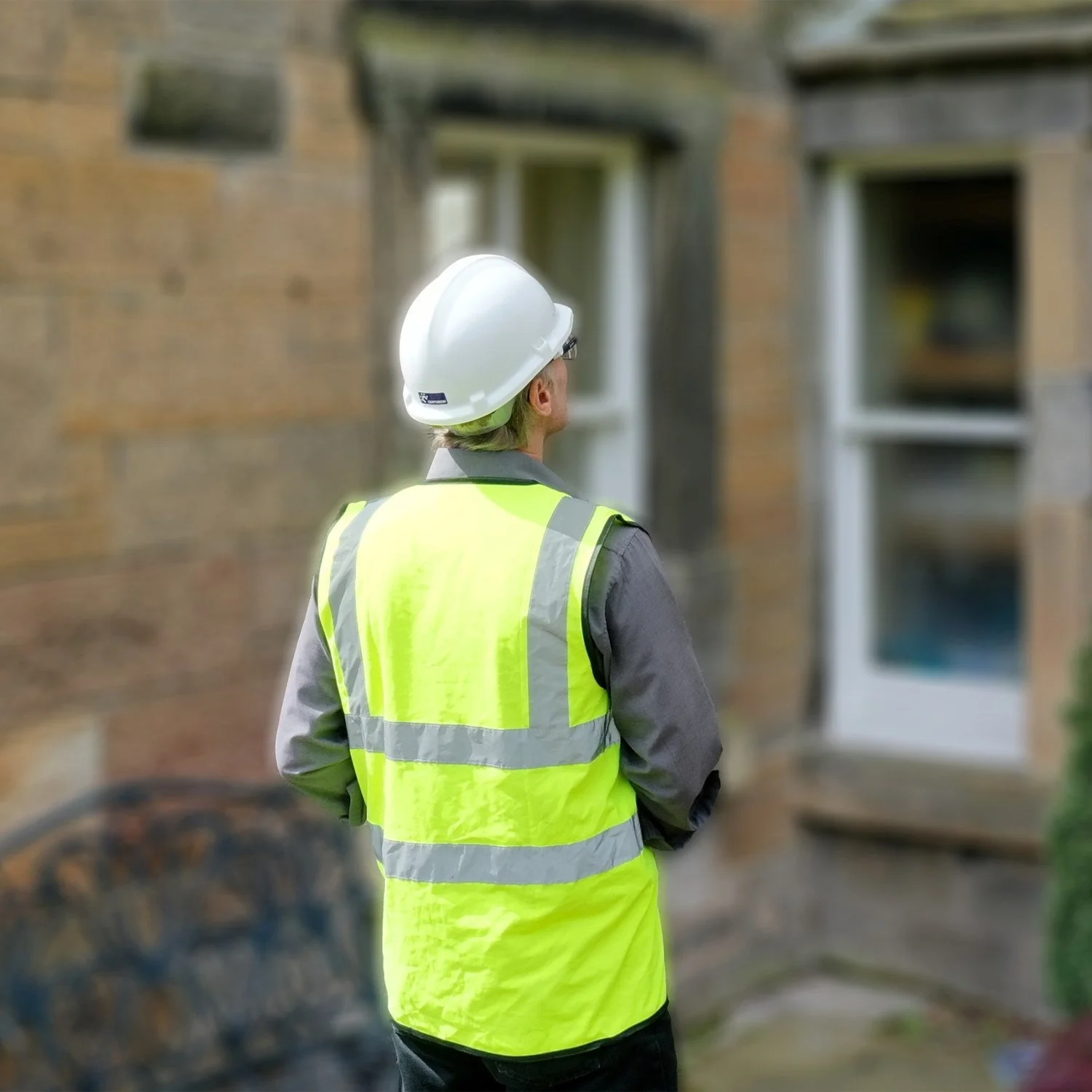 A construction worker wearing a yellow safety vest, a white hard hat, and a gray long-sleeve shirt stands in front of a stone building with windows.