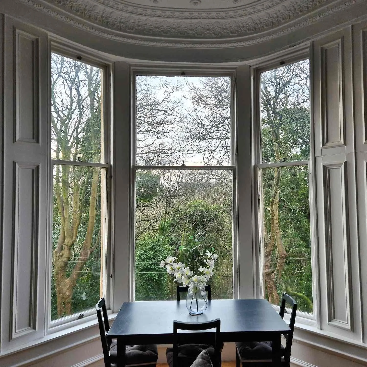 Dining room with a large bay window overlooking trees outside, a table with a vase of white flowers, and black chairs.