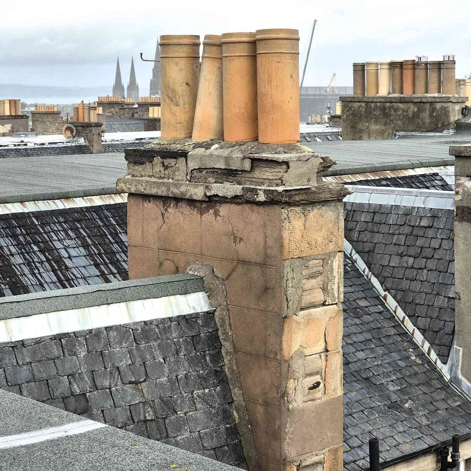 Rooftops of buildings with multiple chimneys, some with weathered stone, and slate roofs, with a cloudy sky and church spires in the background.