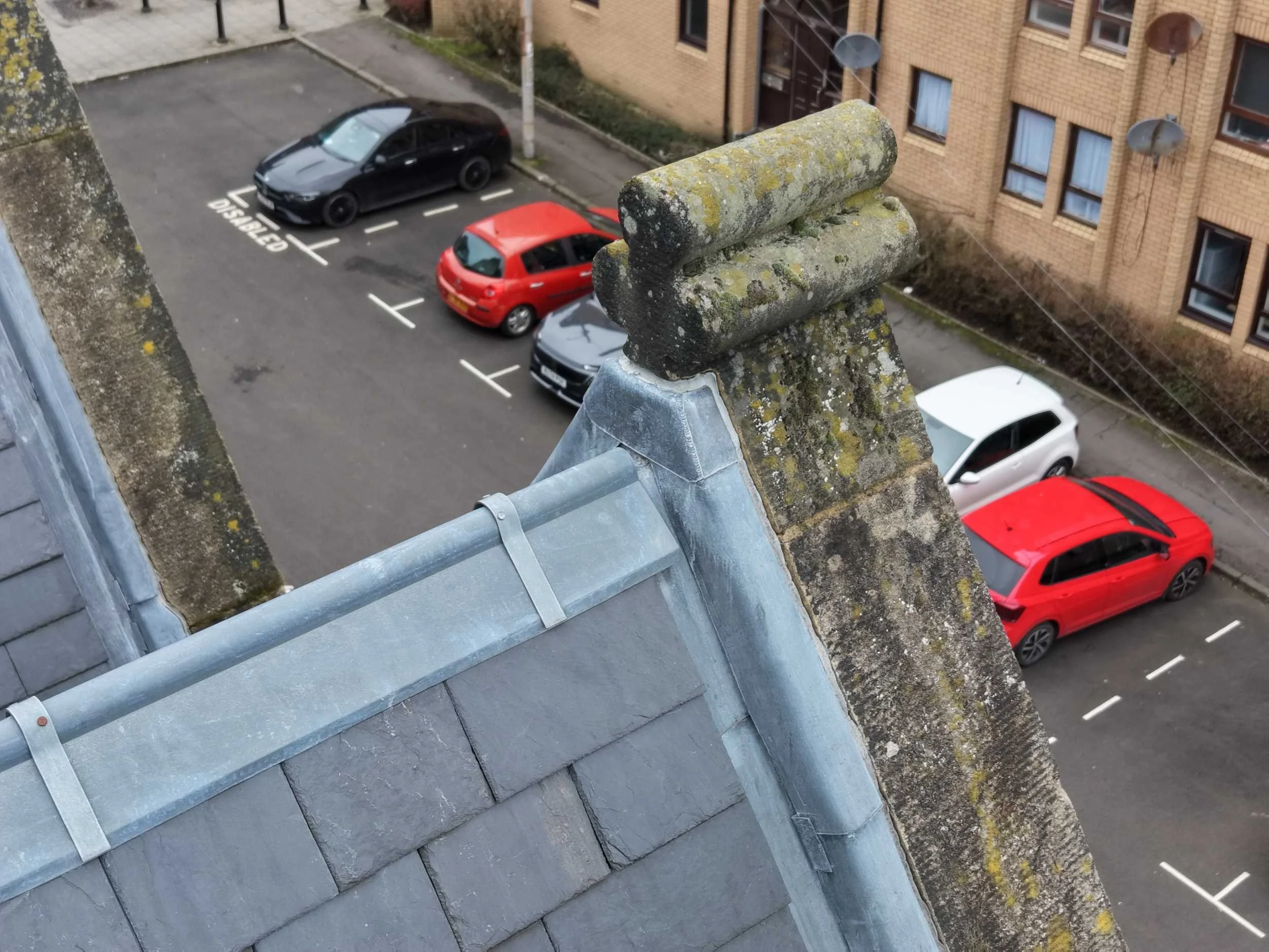 View from a rooftop showing ornate stonework and lead flashing to a slate covered roof, over a street with several parked cars.