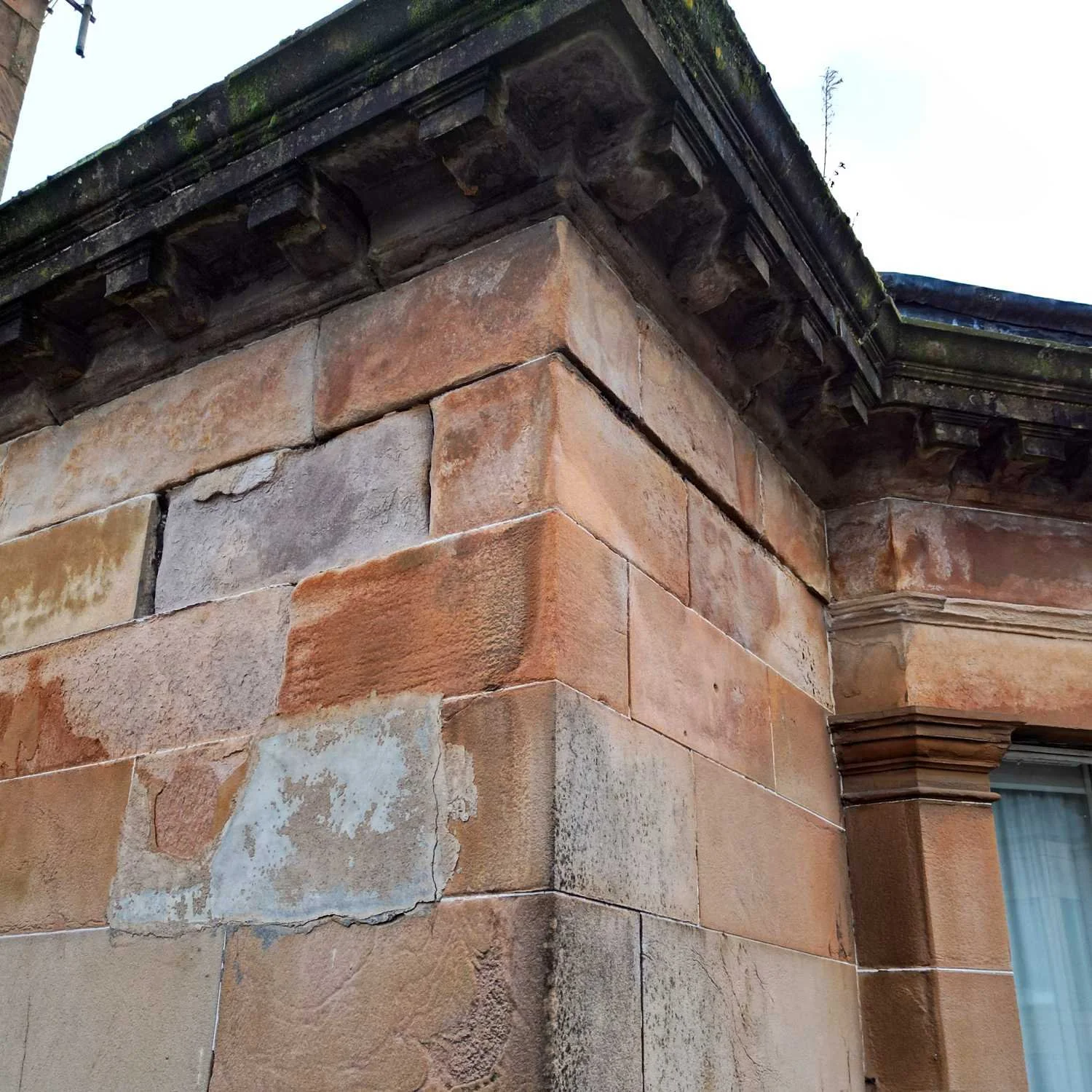 Close-up of the corner of a building with old, weathered stone and a decorative cornice at the top. The stone shows signs of age with varying shades of brown, tan, and gray, and some have visible cracks and patches of peeling paint.