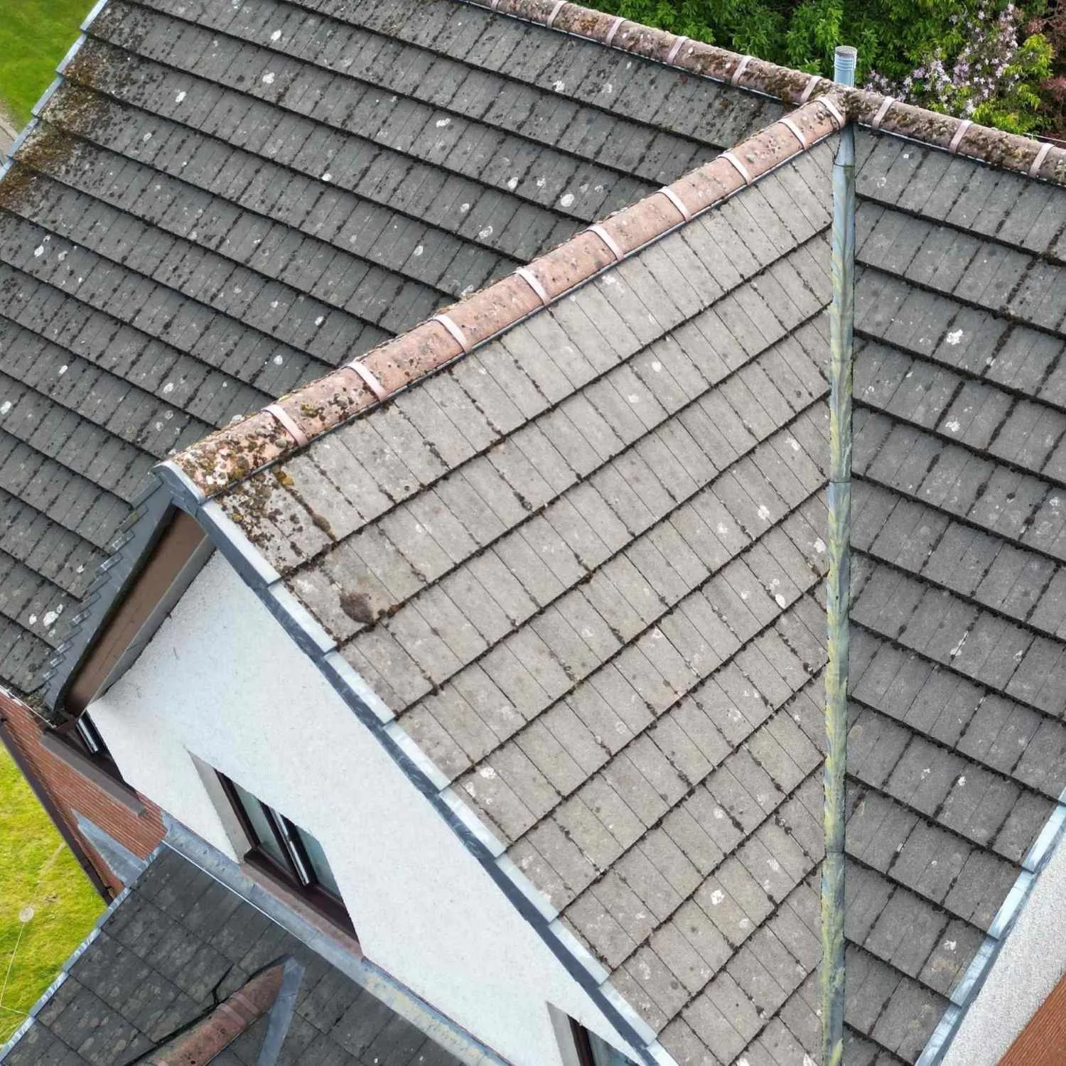 Aerial view of a house roof with weathered gray shingles and moss growth, with a metal chimney pipe running along the roof's ridge, and green trees and a lawn surrounding the house.