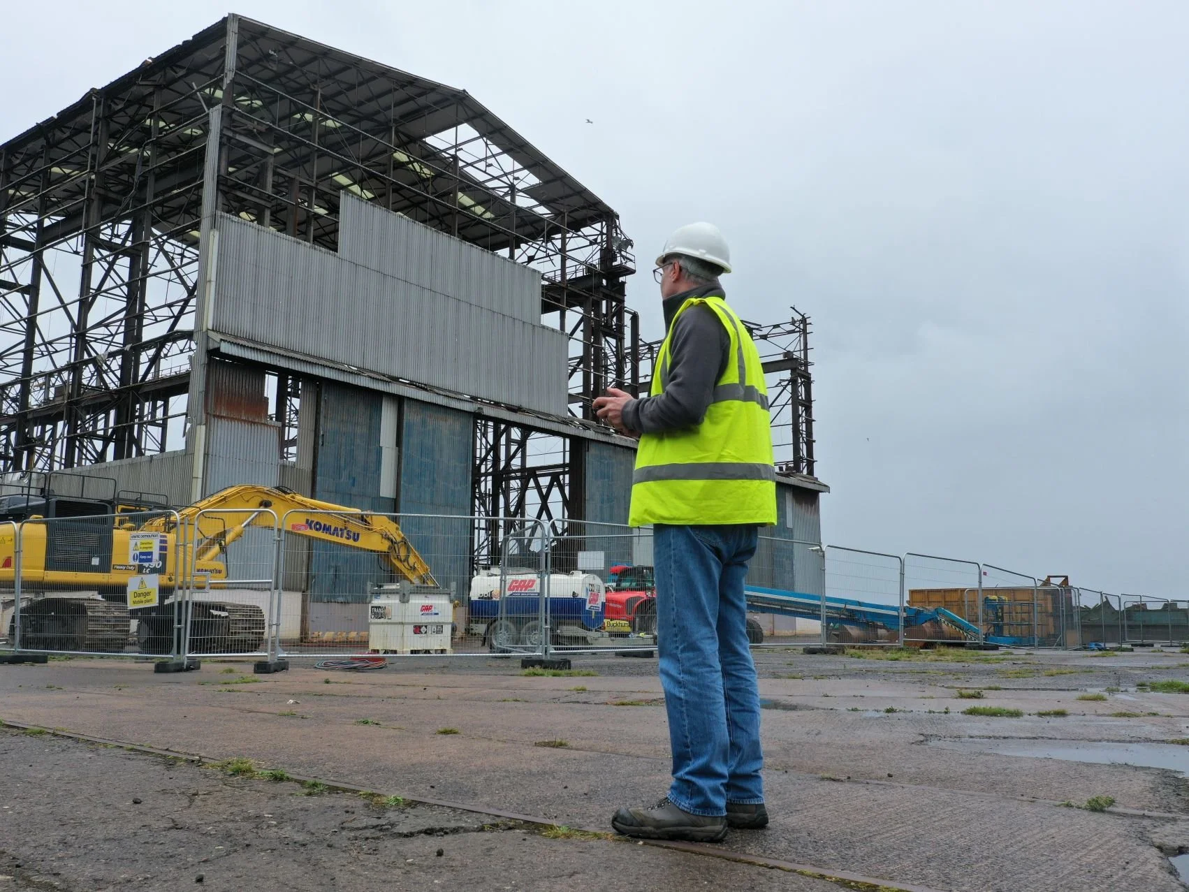 A drone pilot wearing a white hard hat and yellow safety vest, standing outside a construction site with steel framework, construction equipment, and a cloudy sky in the background.