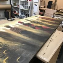 A large, detailed abstract painting on a long, horizontal canvas in a studio, with various colors and textures.