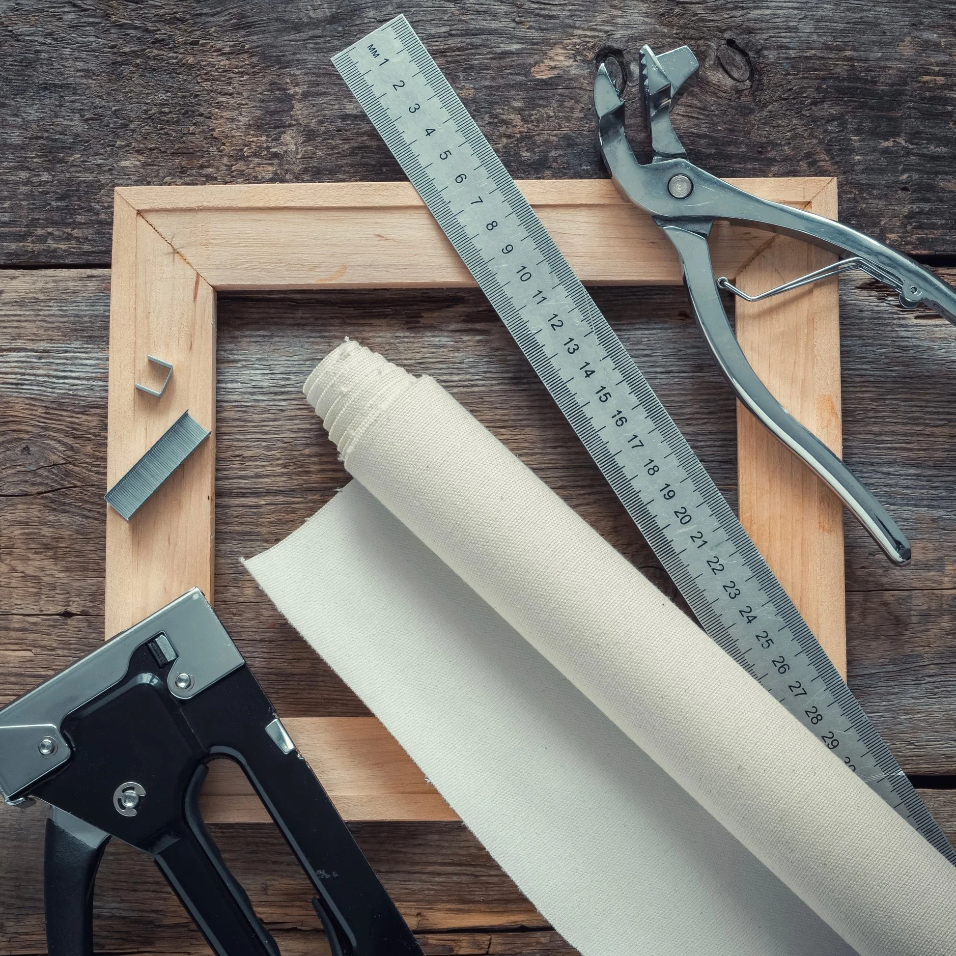 Tools and materials for picture framing, including a wooden picture frame, a metal ruler, a staple gun, a metal clip, a roll of white canvas or fabric, and a pair of staple removers, all arranged on a wooden surface.