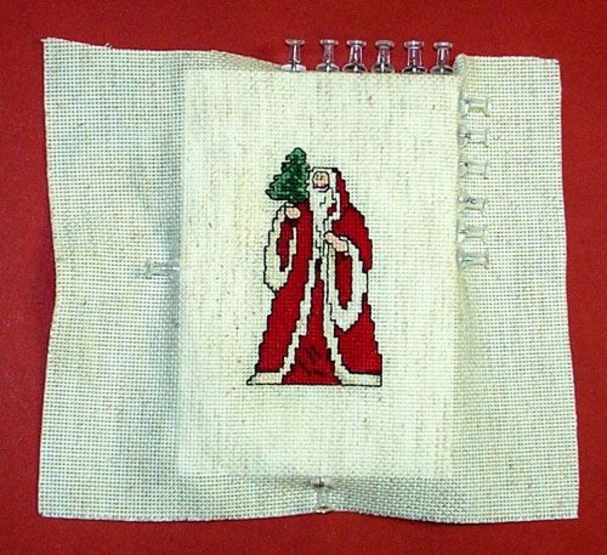 Cross-stitch embroidery of Santa Claus holding a Christmas tree on white fabric, secured with metal clips to a red background.