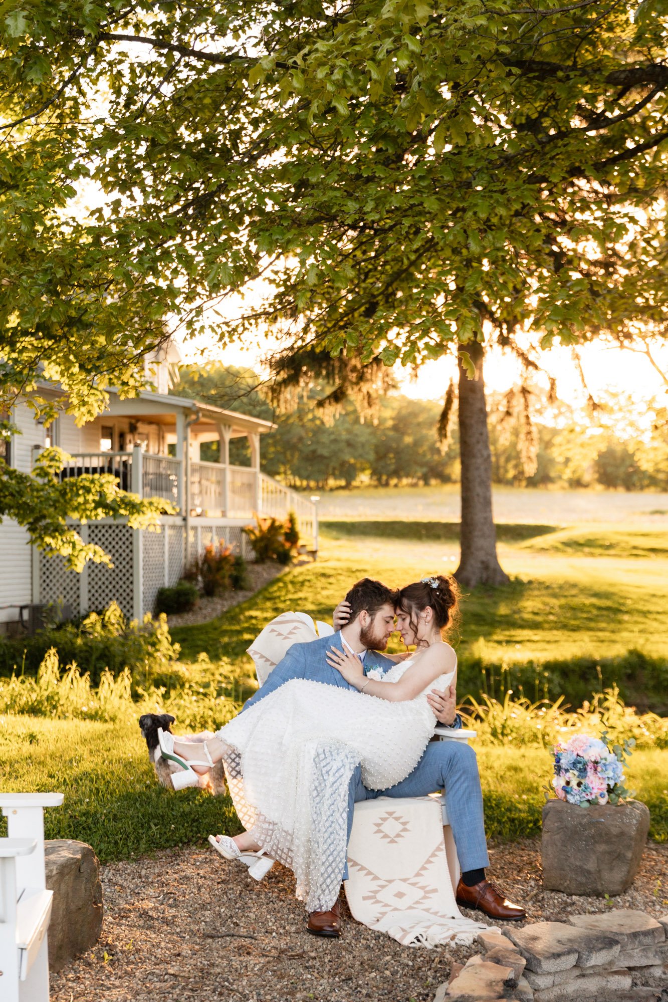 A bride and groom sitting closely together outdoors during sunset, sharing a tender moment with their foreheads touching. The bride is wearing a white wedding dress and heels, and the groom is in a blue suit. A small black and white dog is nearby, an