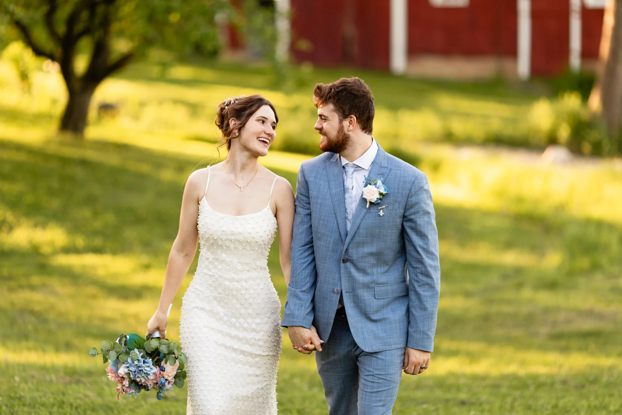 A newlywed couple walking hand in hand outdoors in a park or garden during daytime. The bride is wearing a white dress with a textured pattern, and the groom is in a light blue suit with a boutonnière. They are smiling and looking at each other, hold