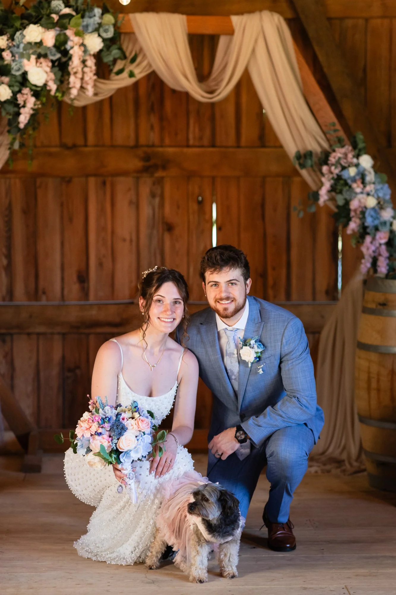 A newlywed couple with a dog in a rustic wedding venue. The bride holds a bouquet of pink, purple, and white flowers, while the groom kneels beside her, smiling. The background features wooden walls with draped fabric and floral arrangements.