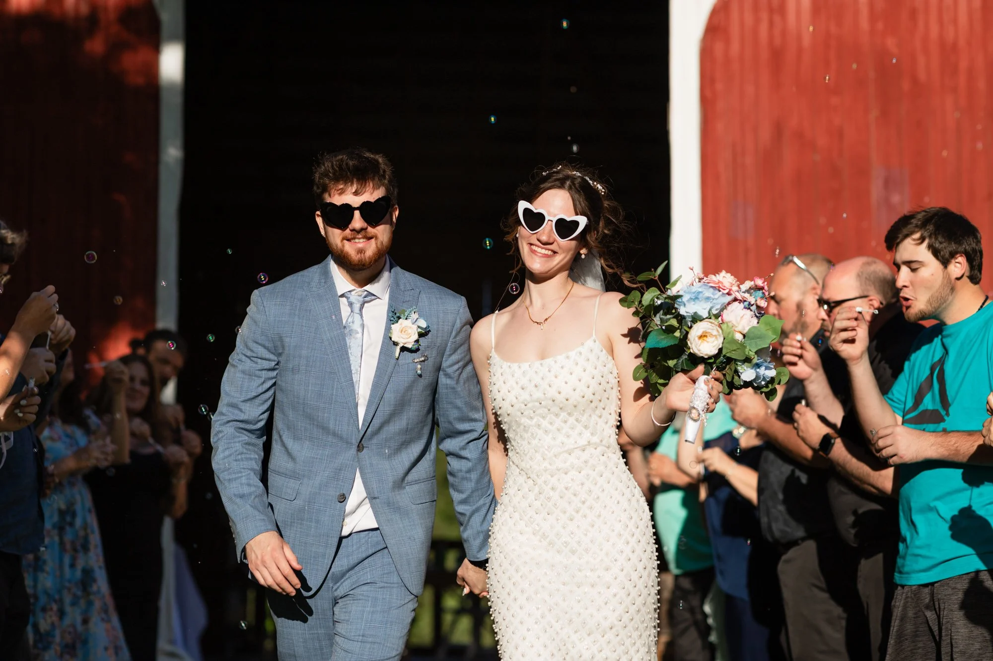 A smiling couple, dressed in wedding attire, holding hands, wearing heart-shaped sunglasses, and walking down an aisle at a wedding ceremony surrounded by guests and bubbles.