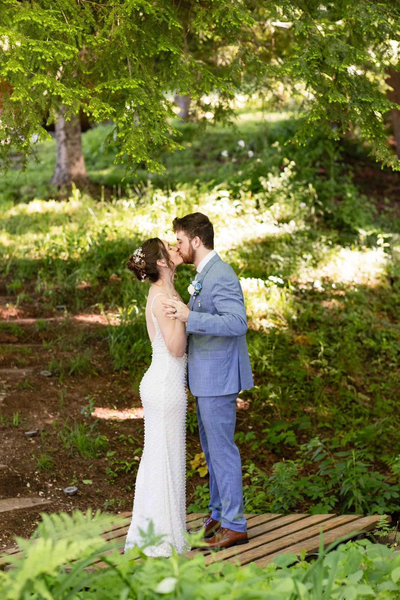 A bride and groom kissing outdoors under green leafy trees, standing on a wooden platform in a garden.