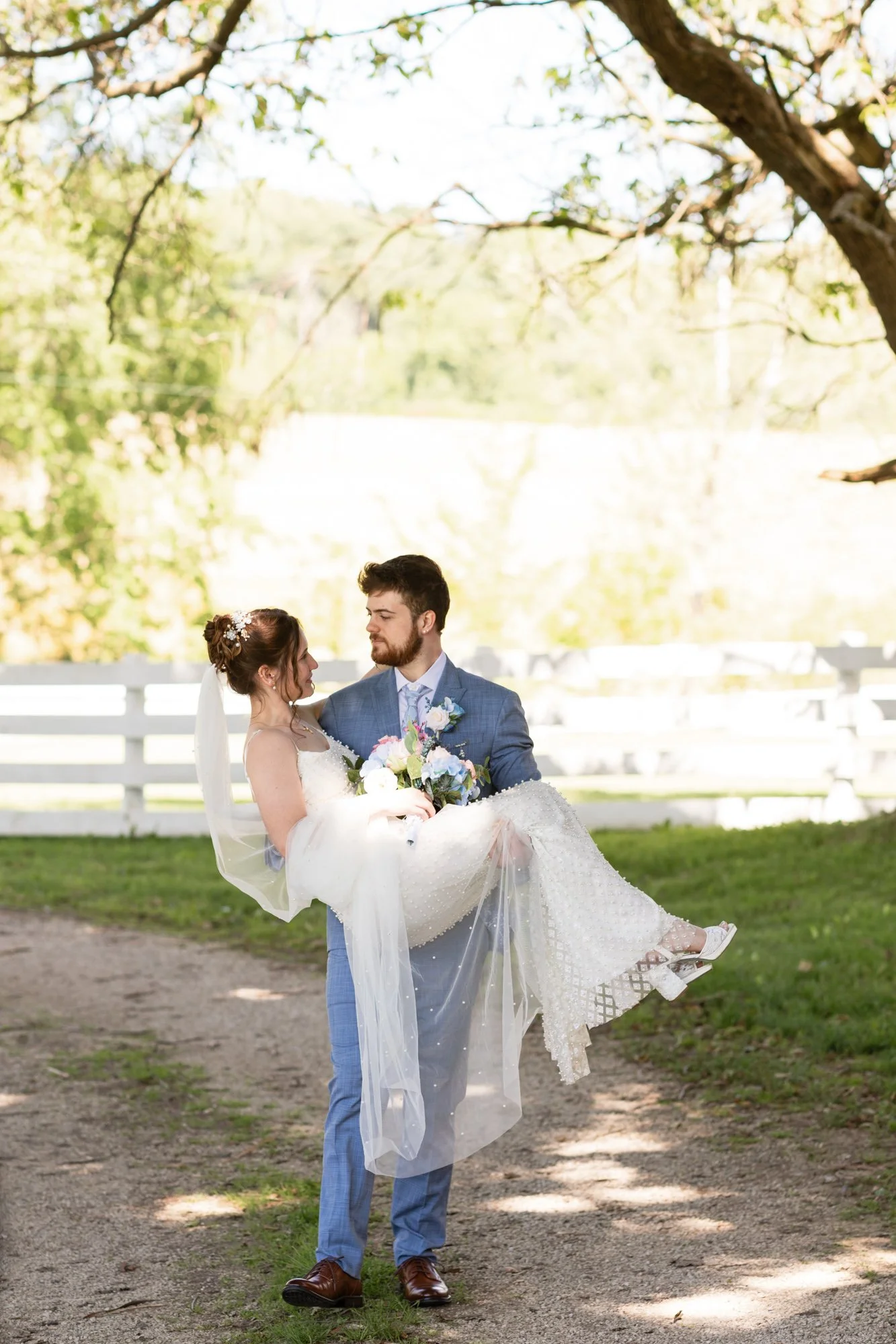 A groom in a blue suit carrying a bride in a white wedding dress outdoors under a tree with a white fence in the background.