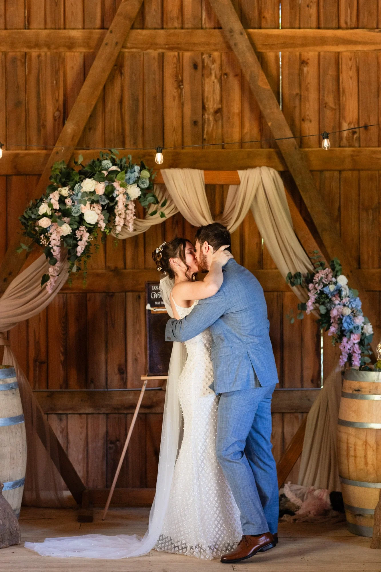 A bride and groom kiss during their wedding ceremony inside a rustic wooden barn decorated with draped fabric and floral arrangements.