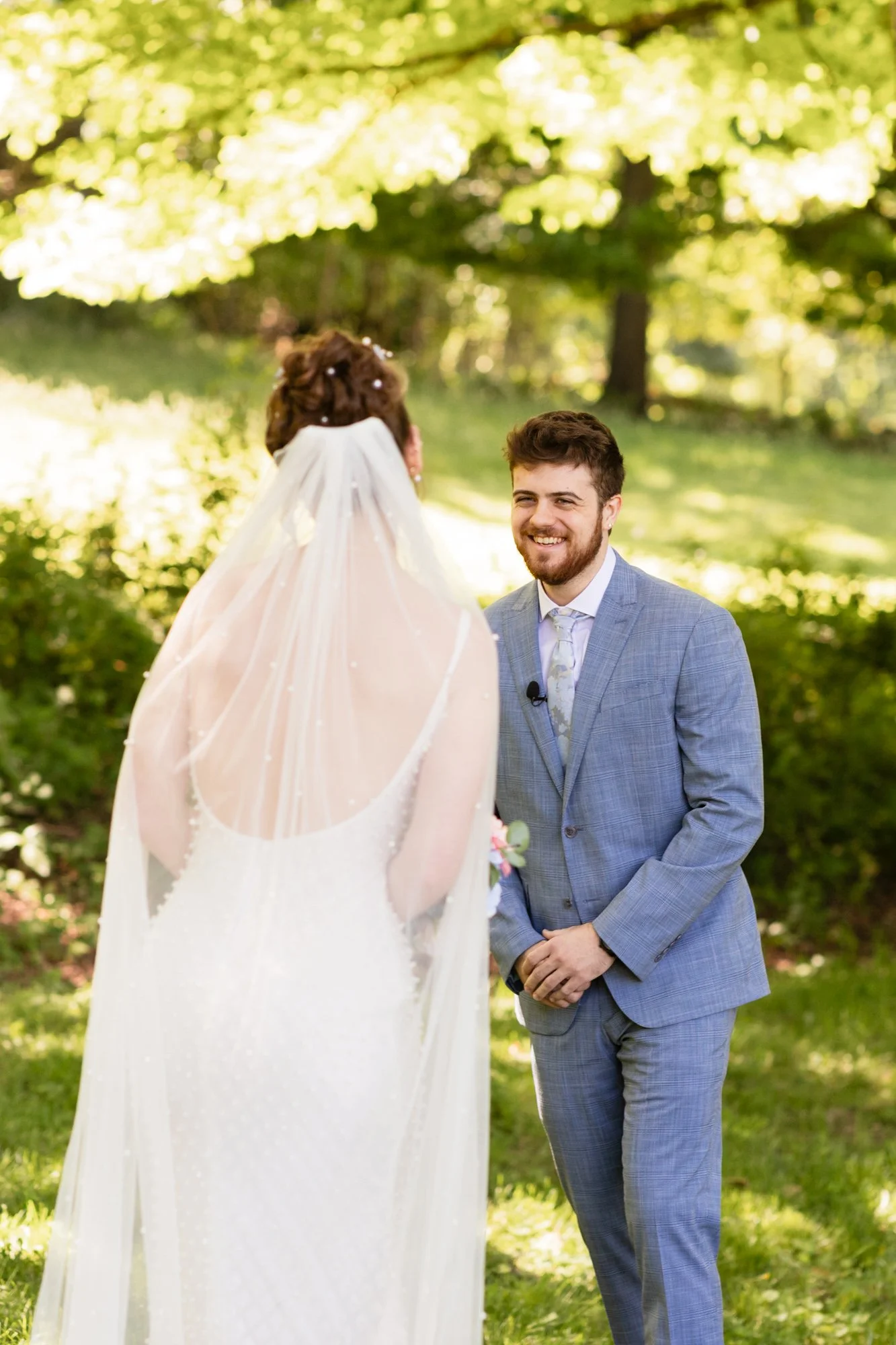 A groom in a light gray suit smiling at a bride in a wedding dress with a veil outdoors with green trees and sunlight in the background.