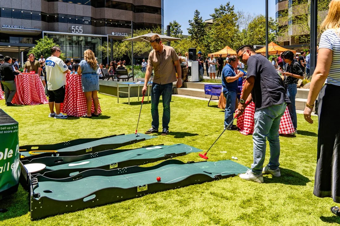 People playing miniature golf on outdoor putting greens during a sunny public event, with food tents and a crowd in the background.