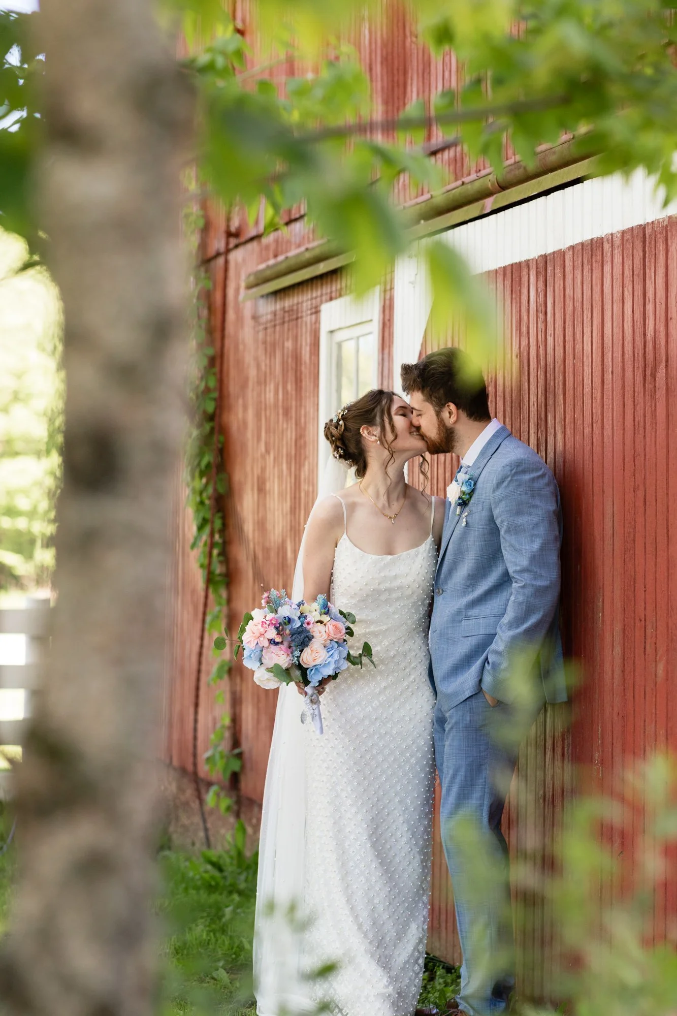 A bride and groom are standing close and about to kiss beside a red wooden building, with greenery and a tree trunk in the foreground.