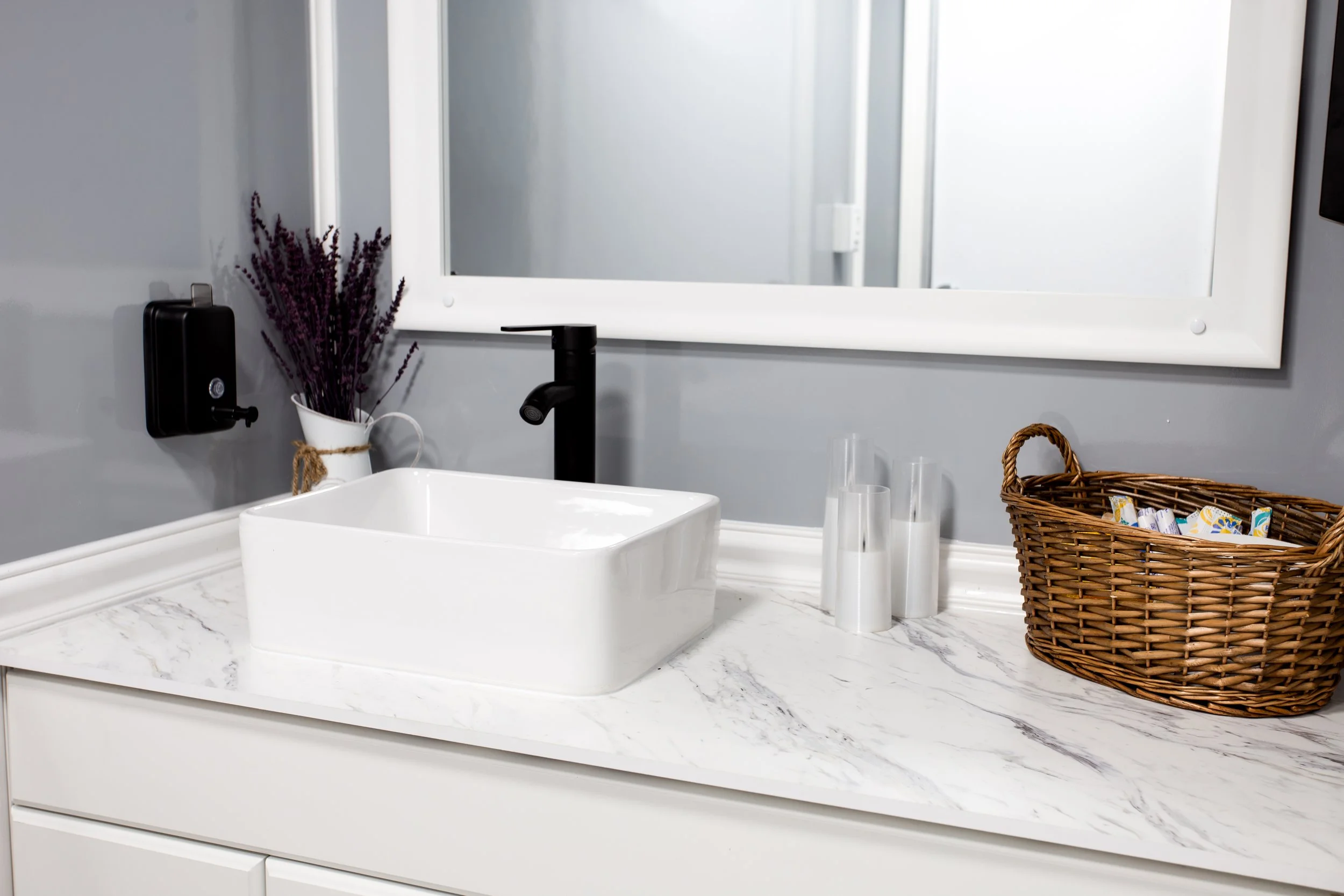 Bathroom sink with a black faucet, a mirror, a vase with purple flowers, a basket with toiletries, and several clear cups on a white marble countertop.