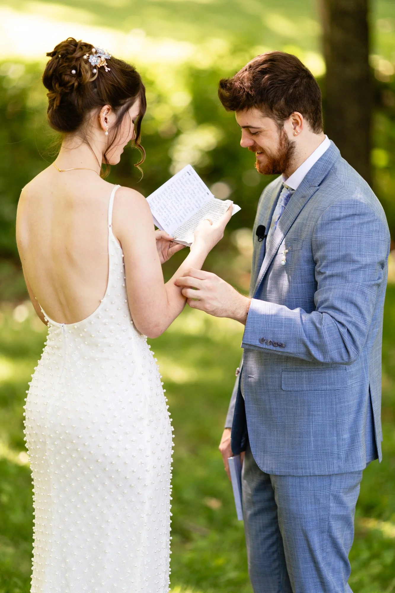 A bride and groom standing outdoors during a wedding ceremony, exchanging vows with the bride holding a piece of paper, both smiling.