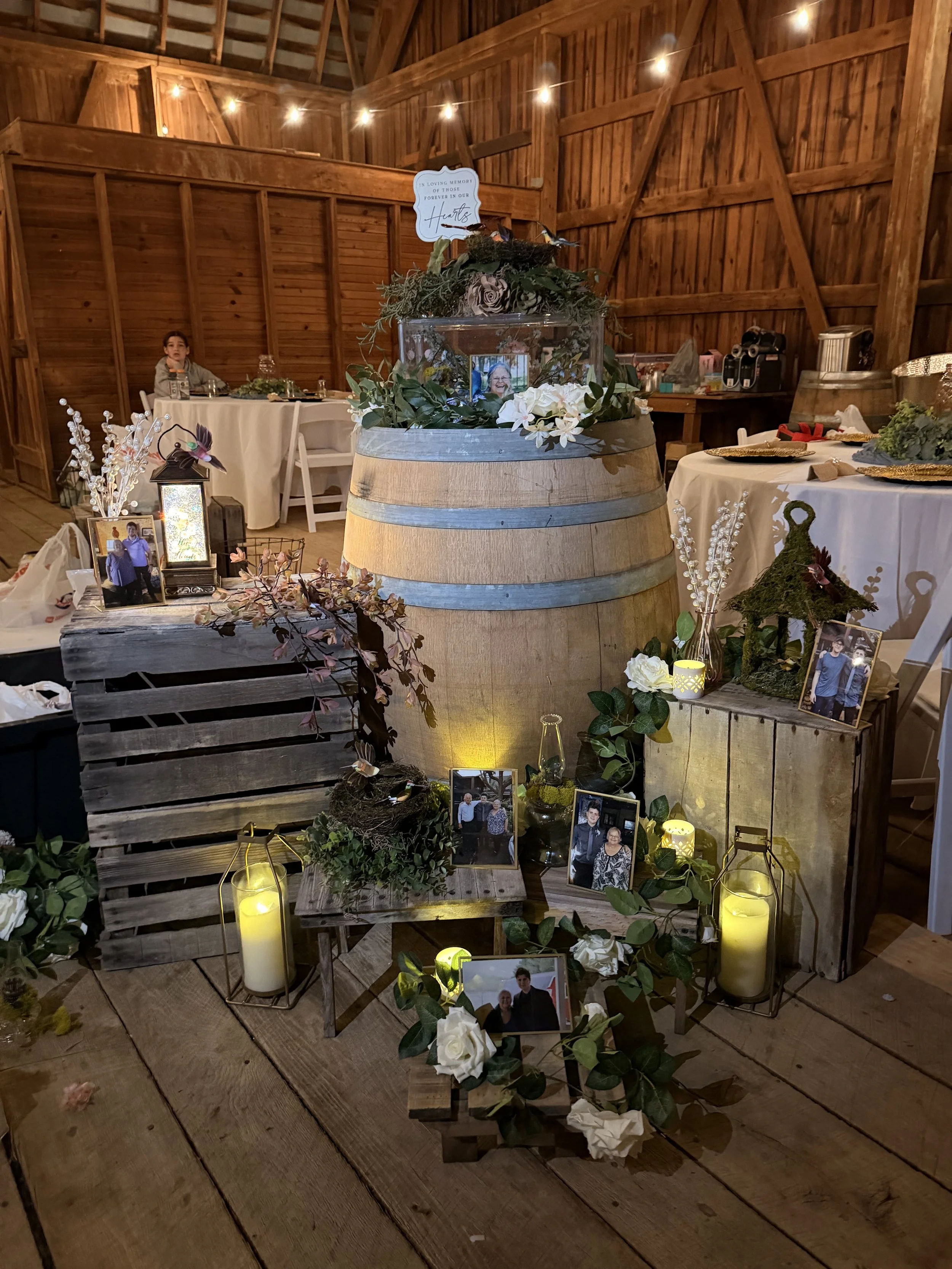 A memorial display with framed photos of loved ones, candles, greenery, and flowers, set in a wooden barn with warm lighting.