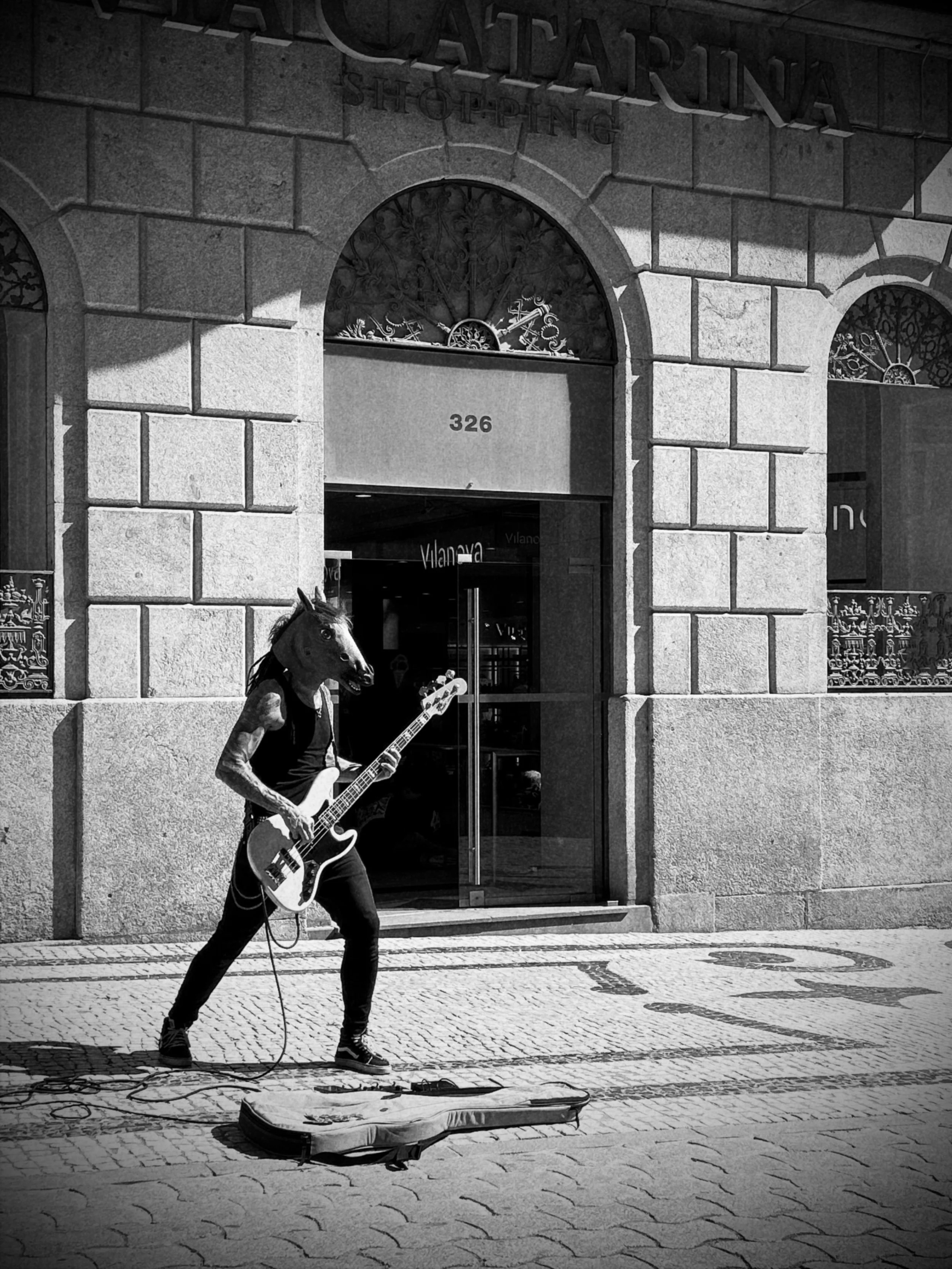 A person dressed as a horse playing a bass guitar on a city sidewalk, with a guitar case on the ground.