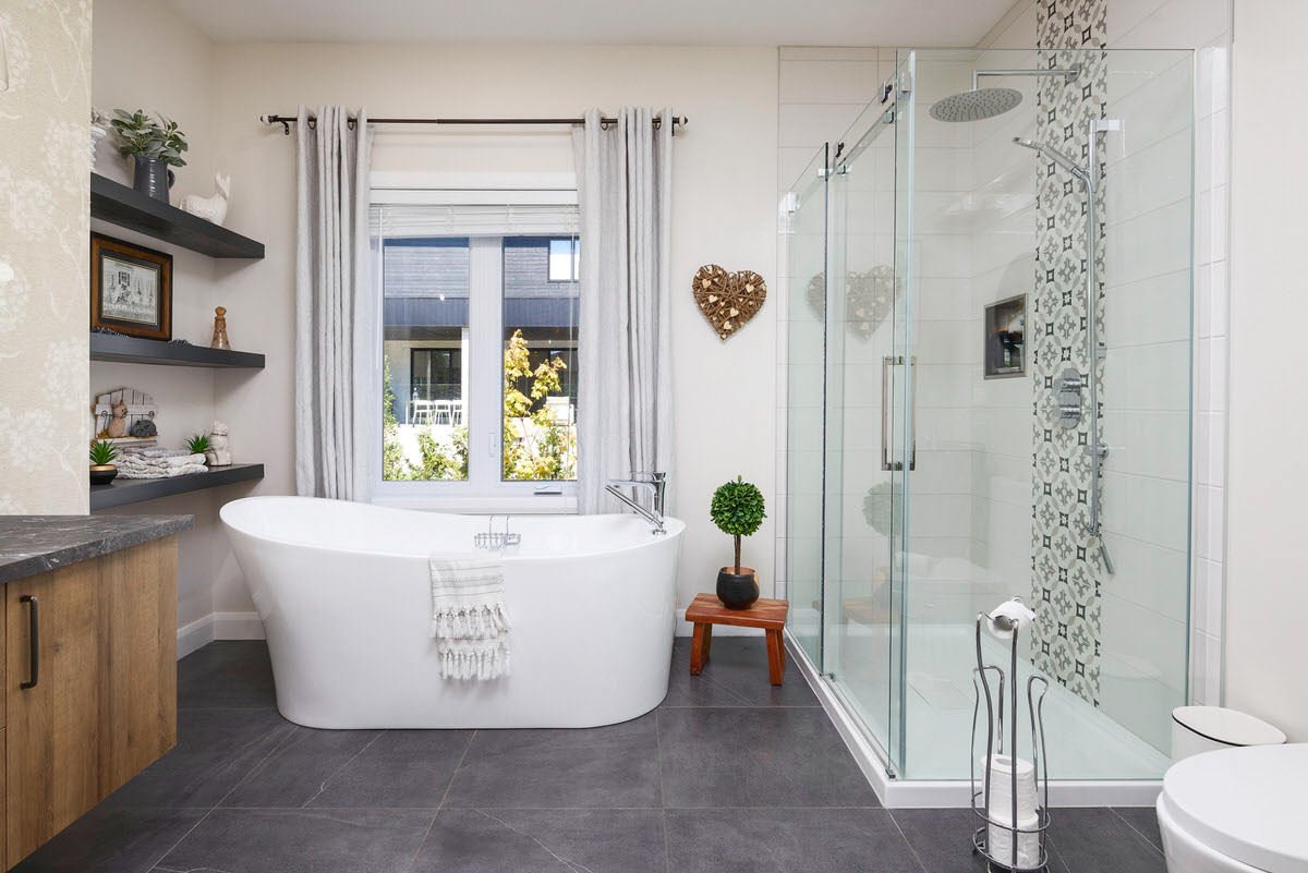 A modern bathroom featuring a white freestanding bathtub near a window with white curtains, a glass-enclosed shower with patterned tile accents, dark gray tile flooring, and decorative shelves with plants and ornaments.