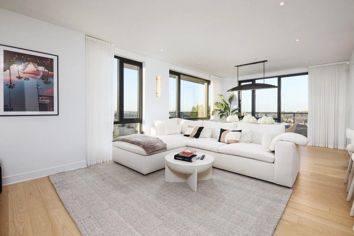 Modern living room with a white sectional sofa, a round coffee table, large windows with white curtains, a framed artwork on the wall, a large plant, and a view of the city skyline.