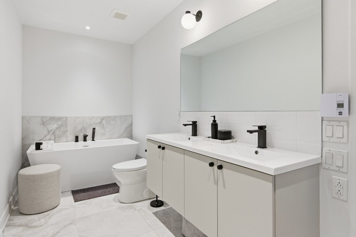 Modern bathroom with white walls, a large mirror, a double black faucet vanity, a bathtub with black fixtures, a toilet, a beige round ottoman, and minimal black accessories.