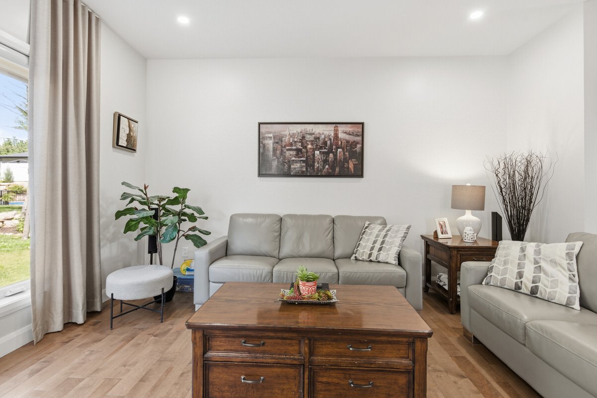 Living room with two light gray sofas, wooden coffee table, side table with lamp, wall art, large plant, window with beige curtains, and decorative pillows.