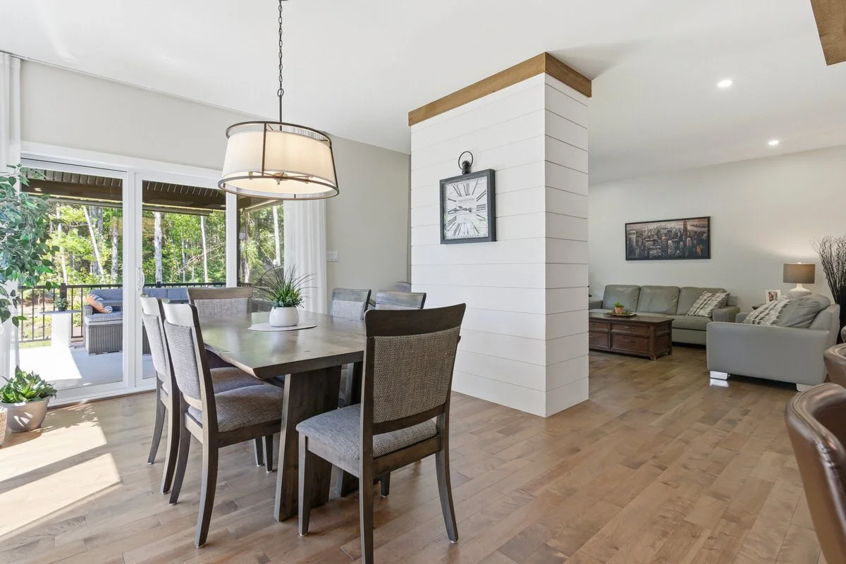 Bright living and dining room with wooden floors, white walls, a dining table with six chairs, a potted plant, and sliding glass door leading to a balcony with outdoor furniture, and the living area with a beige sofa and armchair.