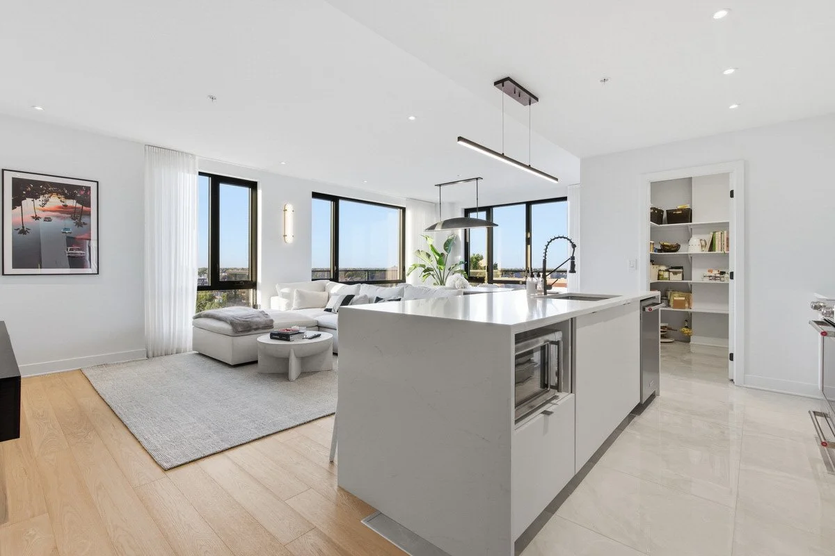 Modern, open-concept living room and kitchen with white walls and cabinetry, hardwood flooring, large windows, and contemporary furniture.