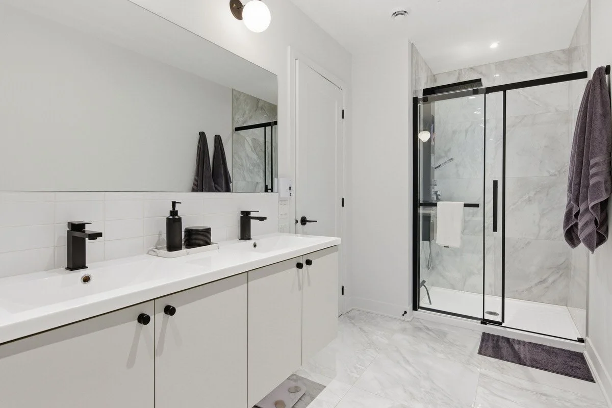 Modern bathroom with double sink vanity, black fixtures, large mirror, and glass shower enclosure with marble walls.