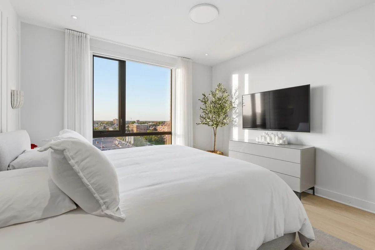 A modern, minimalist bedroom with a large window, white bed with pillows, a flat-screen TV on the wall, and a dresser with candles and a small tree.